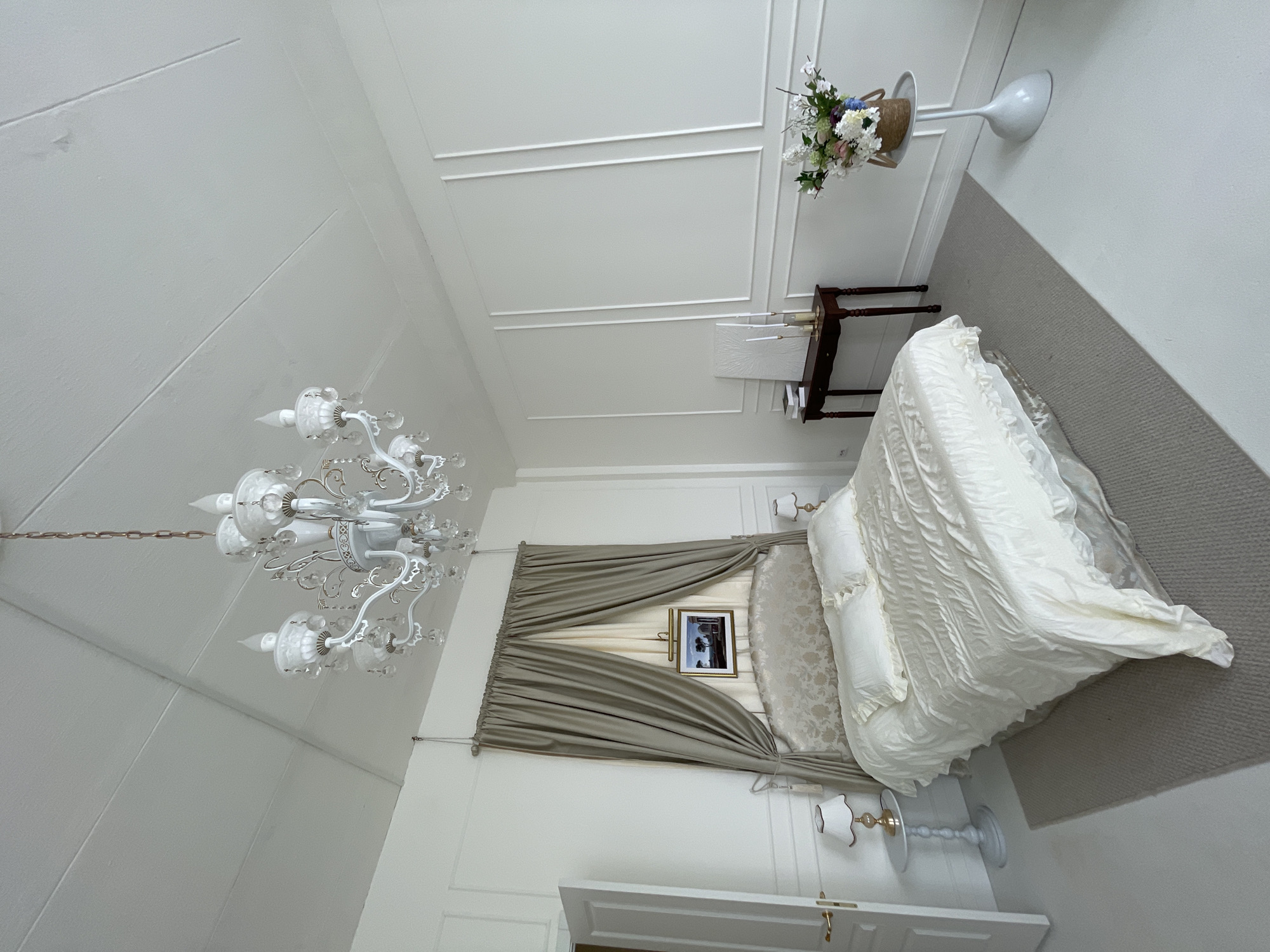 A high-angle, color photograph depicts a bedroom. The room is primarily white, with off-white bedding, a gray carpet, and gray drapes. The bedding covers a bed. The drapes are draped open to either side of a framed picture on the wall above the bed. The room includes a white door at the foot of the bed. There are two small bedside lamps, one on each side of the bed. A small dark wooden table with a lamp sits against the wall above the head of the bed, to the left of a decorative flower display on a white pedestal. A large white chandelier hangs from the ceiling. The photograph was taken in Kazan, Russia.