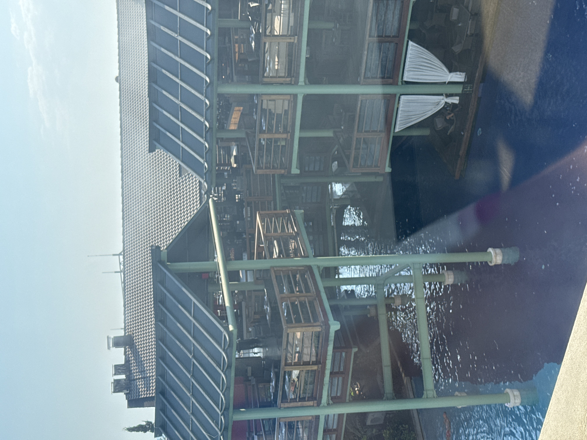 The image depicts a multi-level, open-air restaurant structure built over a body of water. The architecture features dark-colored, sloped roofs with visible beams and supports. The upper levels are open-air dining areas with tables and chairs, enclosed by wooden railings. Some areas have draped white curtains. The structure is supported by thick, light green cylindrical pillars that extend into the water. The water appears to be a pool or a calm waterway with reflections of the structure. In the background, a large, dark-tiled roof with multiple chimneys is visible, suggesting it is part of a larger complex. The sky is clear and bright blue. There are no visible people in the scene. The overall impression is of a resort or dining establishment designed to be integrated with a water feature. This establishment is located in Aksu, Turkey, a coastal district in Antalya.