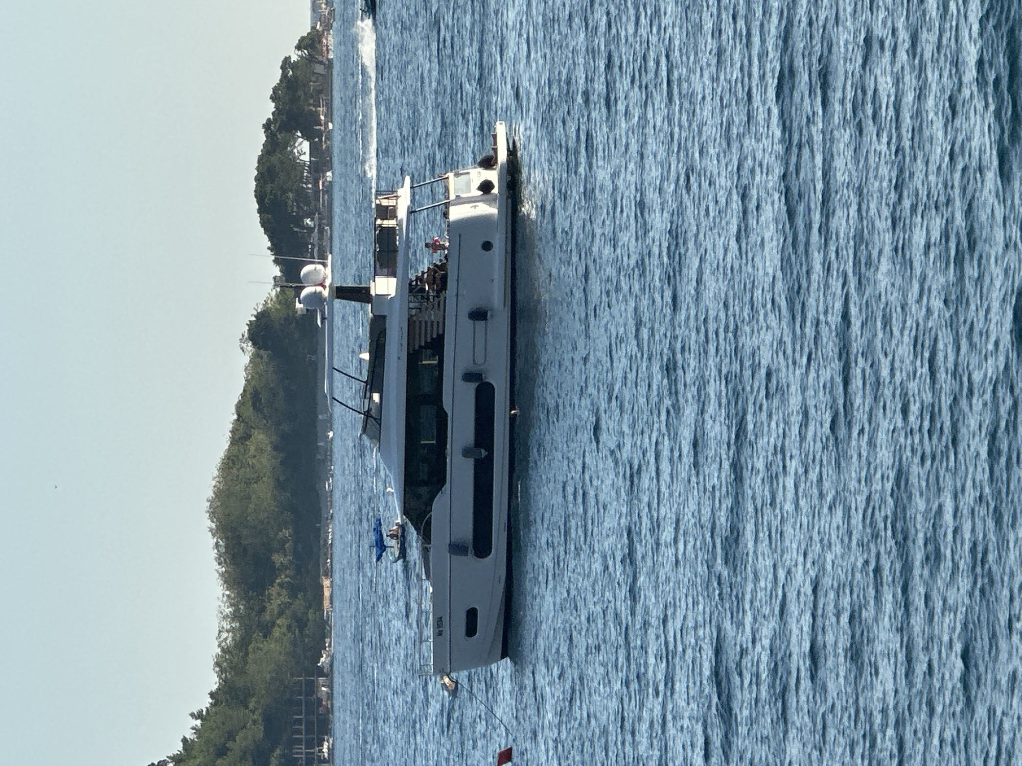 A white motor yacht with a dark hull stripe and a second level deck is docked on blue water. The yacht has a registration number "MS 1034" on its bow. People are visible on the deck of the yacht. In the background, a tree-covered island with buildings and a construction site are visible, along with other boats on the water. The scene appears to be in the city of Budva, Montenegro.