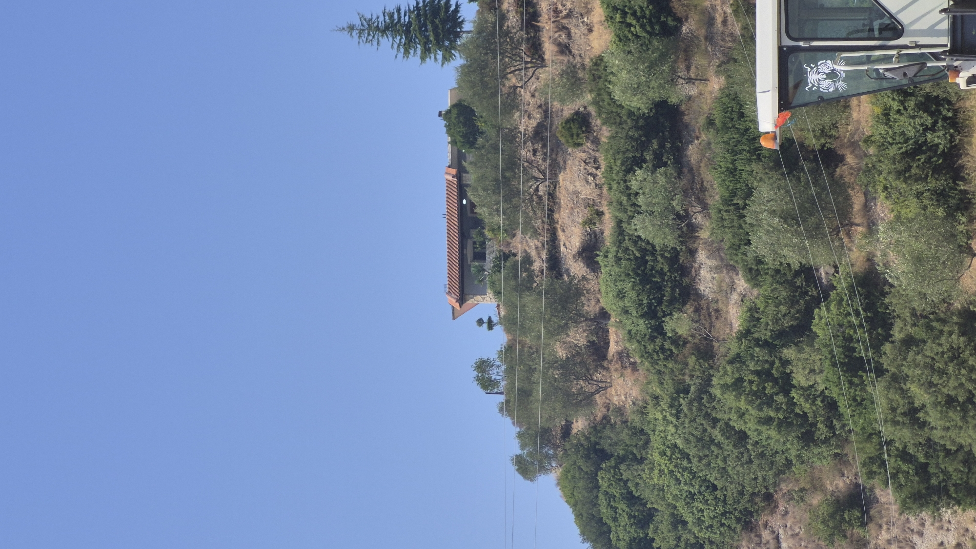 In the city of Hekal, Albania, an outdoor scene unfolds under a clear blue sky. The image captures a hilly terrain dotted with lush green trees and dry, rough ground. Perched atop the hill is a house with a reddish-brown tiled roof and stone walls. Adjacent to the house, a tall, slender evergreen tree stands out. In the foreground, the top of a white vehicle is visible, featuring an orange indicator light and a decorative tiger face decal on its window. Power lines traverse the landscape horizontally. The overall impression is a rural setting with a dwelling on a hillside, possibly a farm or a remote residence.