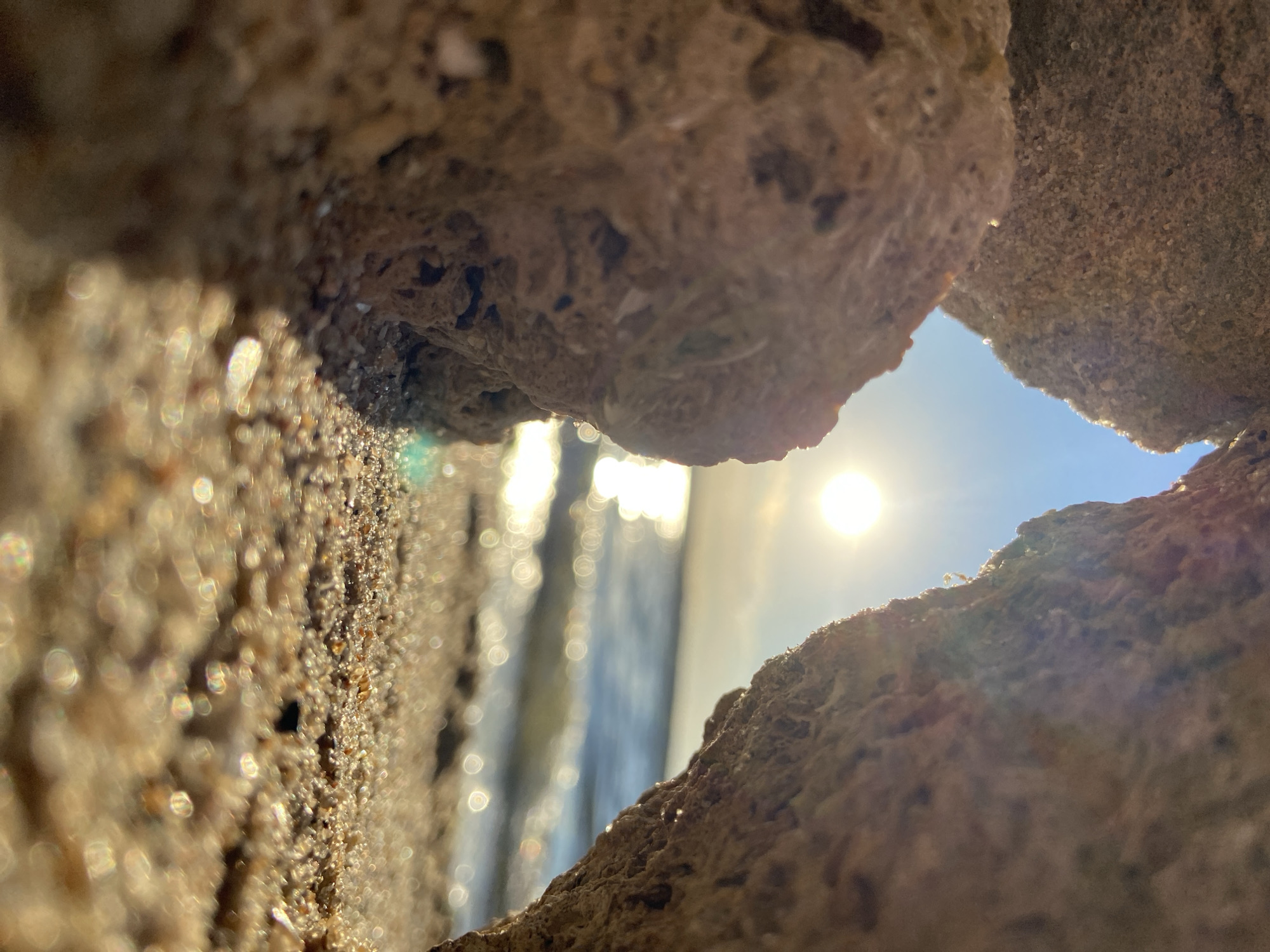 The image is a close-up, low-angle shot looking through a gap between two large, textured rocks. The rocks appear to be rough and porous, with a sandy or granular surface. The gap between them frames a view of the ocean and the sky. The sun is visible in the sky, shining brightly and causing a glare. There are also visible water droplets or reflections on the rocks and in the foreground, creating a bokeh effect. The overall scene suggests an outdoor setting, possibly a beach or rocky coastline, with bright daylight. The focus is on the texture of the rocks and the light filtering through the opening, creating an abstract and somewhat ethereal feel. There is no discernible text in the image.
