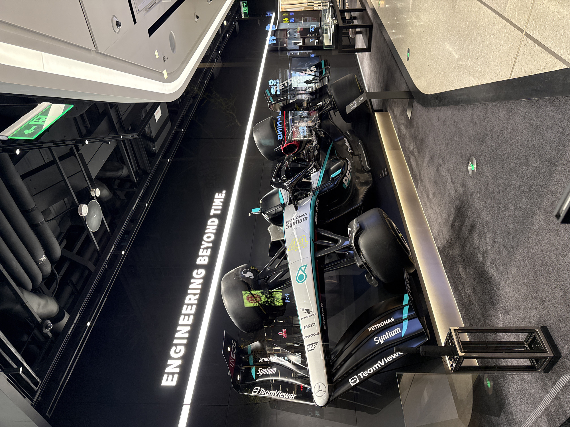 The image shows a Mercedes-AMG Petronas Formula One Team car on display, likely in a retail or exhibition space. The car features prominent sponsor logos such as Petronas, TeamViewer, and Ineos. Text on the wall reads "ENGINEERING BEYOND TIME". The environment includes a black wall and ceiling with visible piping and lighting fixtures. There are at least three race cars on display. It is not possible to confirm from the image if this scene is in Dongcheng District, China.