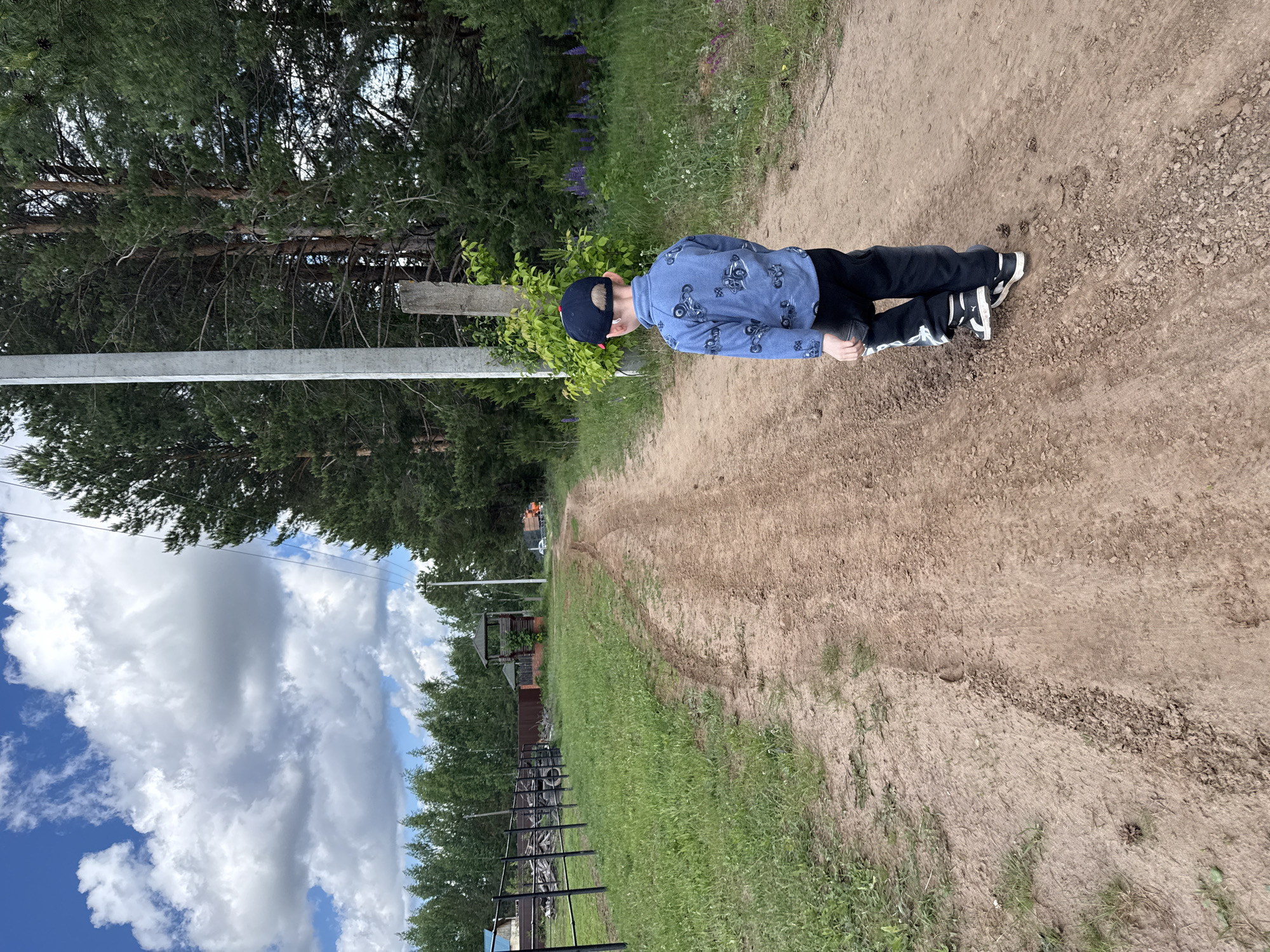 A young boy wearing a blue motorcycle-print shirt, black pants with white stripes, and a dark baseball cap is lying face down on a dirt path. The path is flanked by green grass and trees. There's a wooden post and an overhead metal support beam to the left. In the background, a fence runs along the edge of the grass. Above, there are cumulus clouds and blue sky. The location is likely the city of Chaikovsky, Russia.