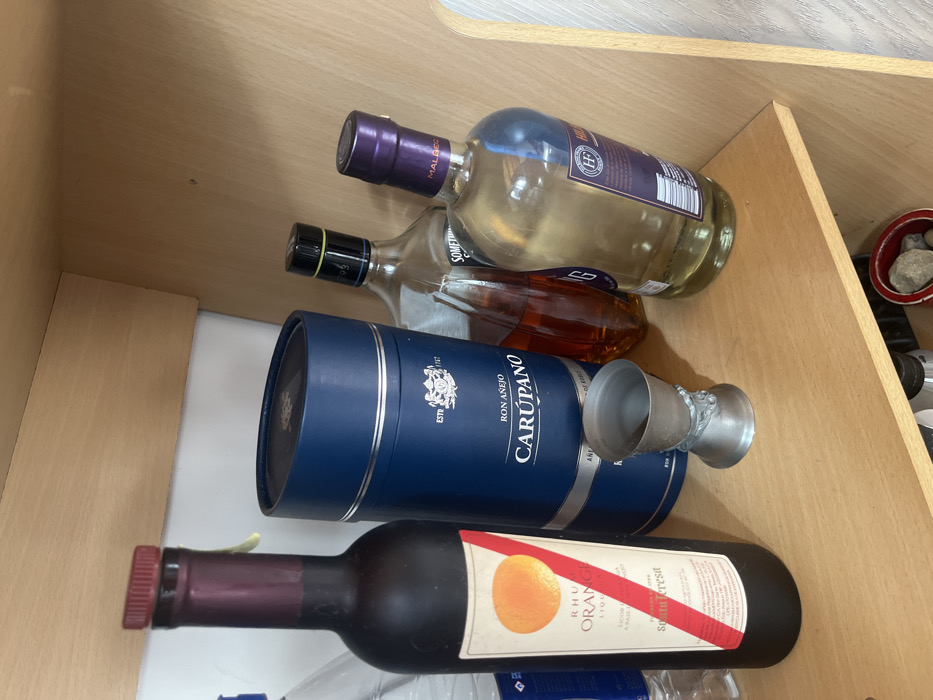 The image displays an interior scene within a wooden storage unit or cabinet, located in Ushuaia, Argentina. A collection of alcoholic beverages and a single measuring jigger are arranged on a light-colored wooden shelf. On the far left, a clear plastic bottle is partially visible. Next to it stands a dark brown glass bottle of Santa Teresa Rhum Orange Liqueur, featuring an orange circle graphic and a red diagonal stripe on its label. Adjacent to the liqueur bottle is a dark blue cylindrical presentation box for Ron Añejo Carúpano, marked with the brand name, "RON AÑEJO," and "ESTD 1767." In front of the Carúpano box, a metallic double jigger, with a detailed decorative base, rests on the shelf. To the right, a clear glass bottle filled with an amber-colored spirit is present, featuring a black cap with a yellow band. Beside it is another clear glass bottle containing a pale yellow liquid, identifiable by a purple neck wrap labeled "MALBEC" and an "HP" logo on its main label. The background behind the shelf is a plain white surface. A small, red, circular container holding several grey rocks is partially visible on a lower shelf in the bottom right corner of the frame.