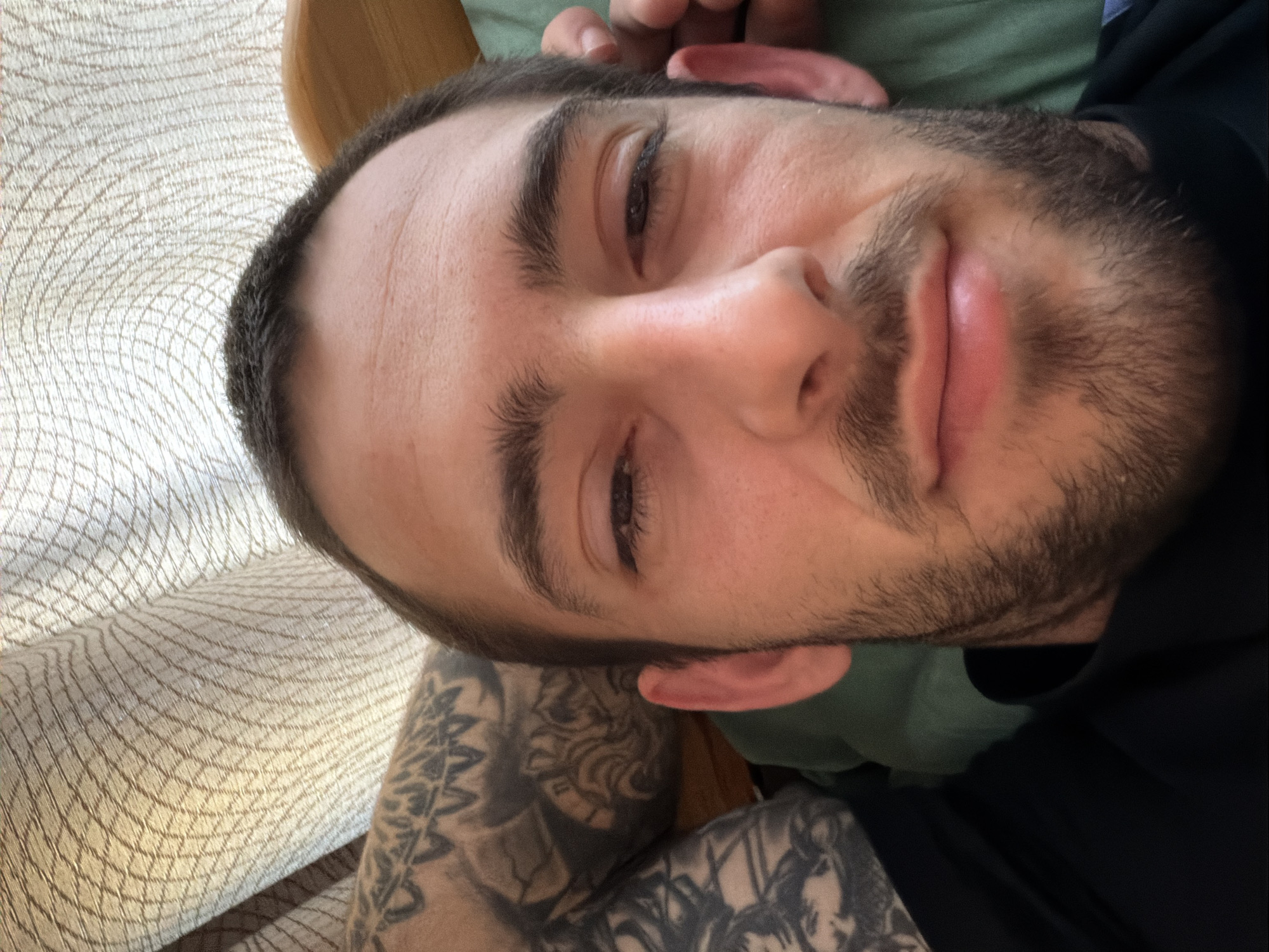 A close-up selfie of a man with dark hair and a beard, lying on what appears to be a chair or sofa with a patterned cream-colored fabric. The man has tattoos on his left arm. He is wearing a dark-colored shirt or top. There is green fabric visible behind his head. The location is unknown and cannot be determined as Plovdiv, Bulgaria.