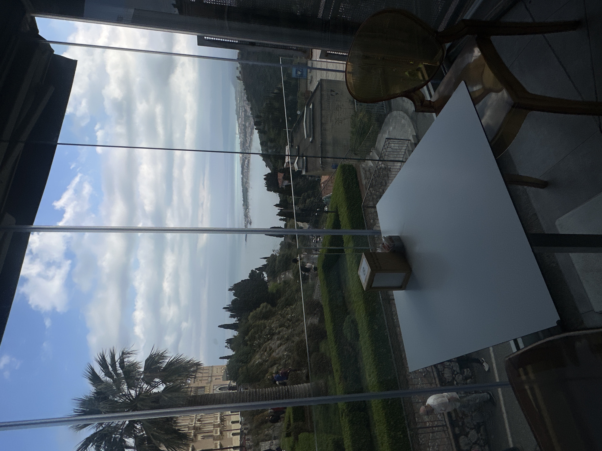 The image shows a view of Taormina, Italy, through a window. A white table with a napkin holder and two chairs are visible inside, close to the window. Outside, there is a park with trimmed bushes and trees, leading towards the coastline and the sea. In the foreground, there is a palm tree. A building with arches is visible in the distance. People are walking on a path adjacent to the park. The sky is partly cloudy. The image seems to be taken from inside a restaurant or building with a view.