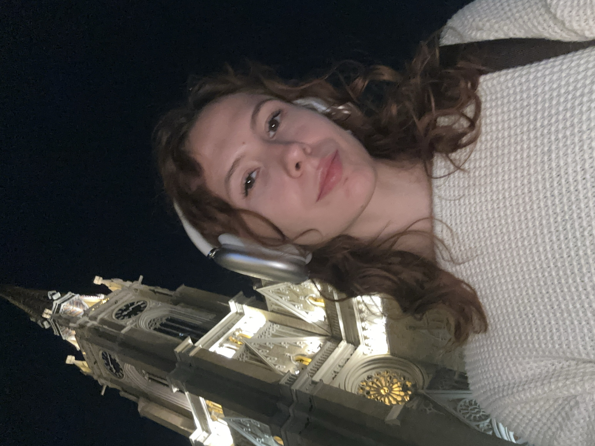 A woman with curly brown hair and wearing white headphones and a light-colored sweater is taking a selfie at night. Behind her is the illuminated facade of the Name of Mary Church, a Roman Catholic church located in Novi Sad, Serbia. The church is characterized by its Gothic Revival architecture, including a tall spire, pointed arches, and intricate stonework. The sky is dark.