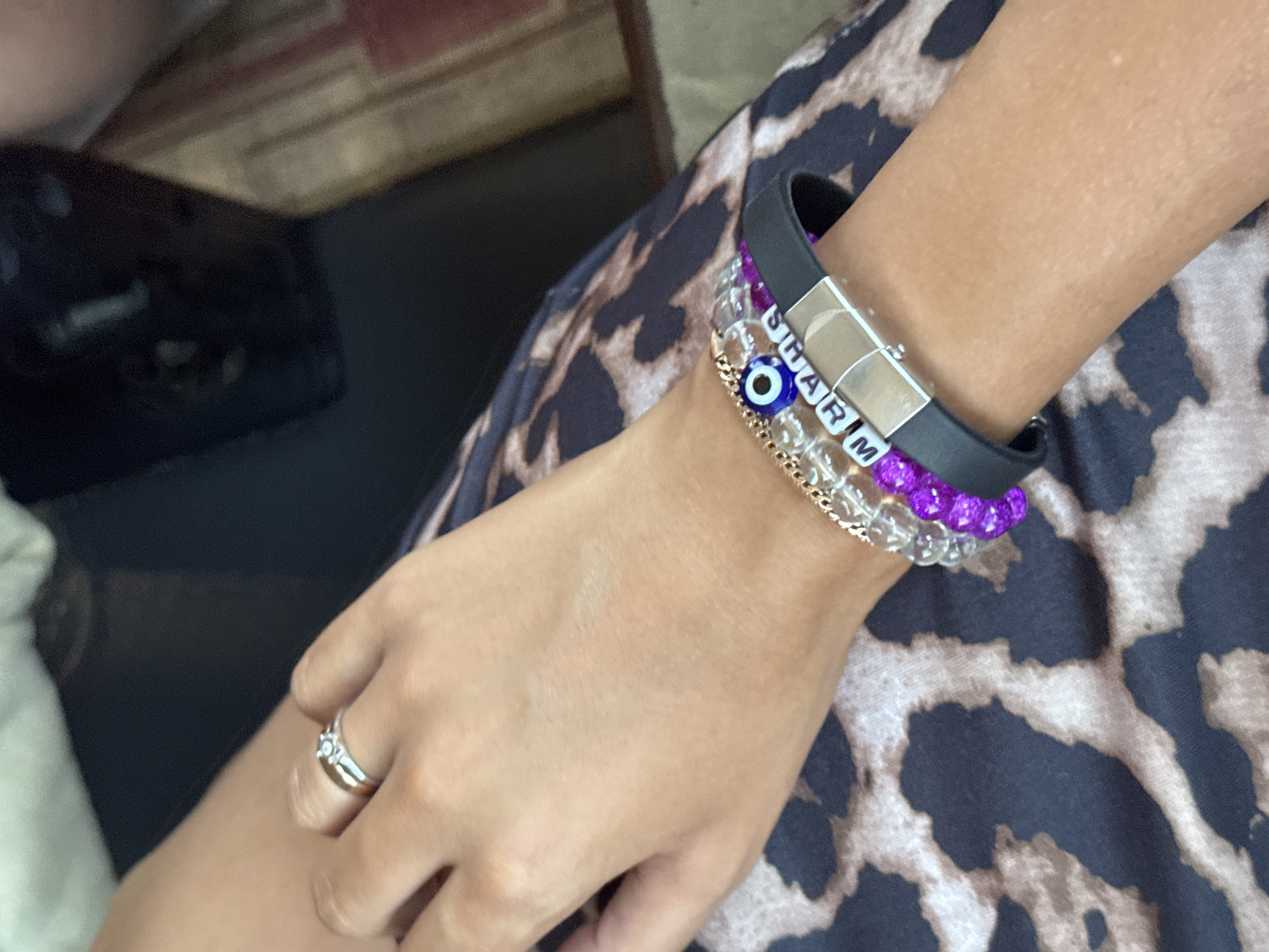 A close-up shot focuses on a person's arm and hand, adorned with several bracelets. One bracelet features white square beads spelling out "SHARM," a blue and white evil eye charm, and clear beads. Another bracelet is made of purple beads, and a third is a black band with a silver clasp. A silver ring with a small stone is visible on the person's finger. The background shows a dark surface and a leopard-print fabric, possibly clothing. There is no direct visual information in the image that places it specifically in Sharm El Sheikh, Egypt, beyond the inscription on one of the bracelets.