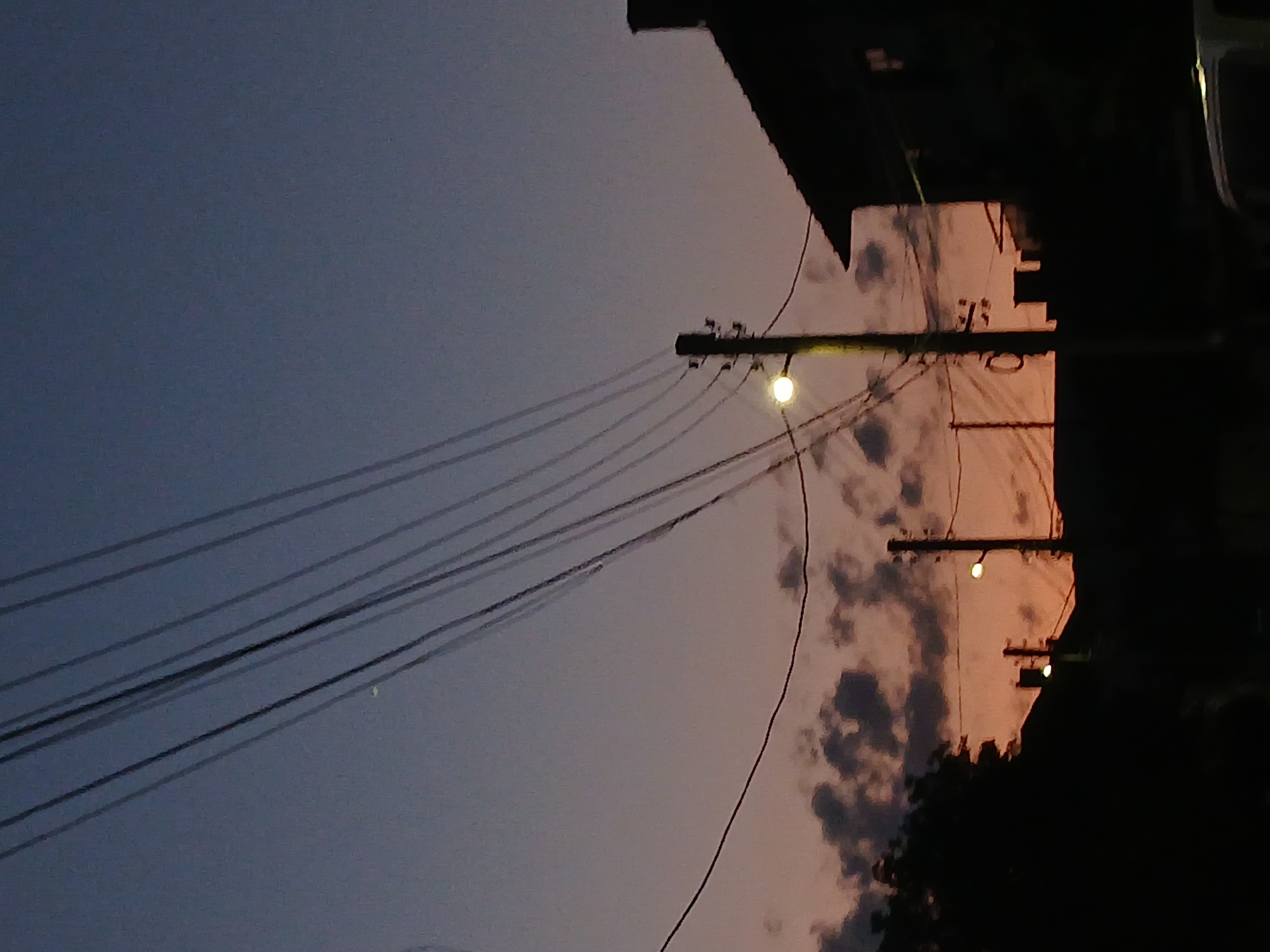 The image depicts a dimly lit outdoor scene, likely during twilight, in Panagyurishte, Bulgaria. The sky transitions from a deep blue at the top to a soft orange-pink hue near the horizon, with scattered dark clouds visible. Several electrical wires stretch diagonally across the frame, dominating the upper portion of the image. A wooden utility pole with a glowing street lamp stands prominently in the mid-ground. To the right, the silhouette of a building's roofline is visible against the sky. The foreground is almost entirely black, obscuring details of the ground and any potential lower structures or vehicles. The overall impression is one of a quiet evening in a residential or semi-urban area.