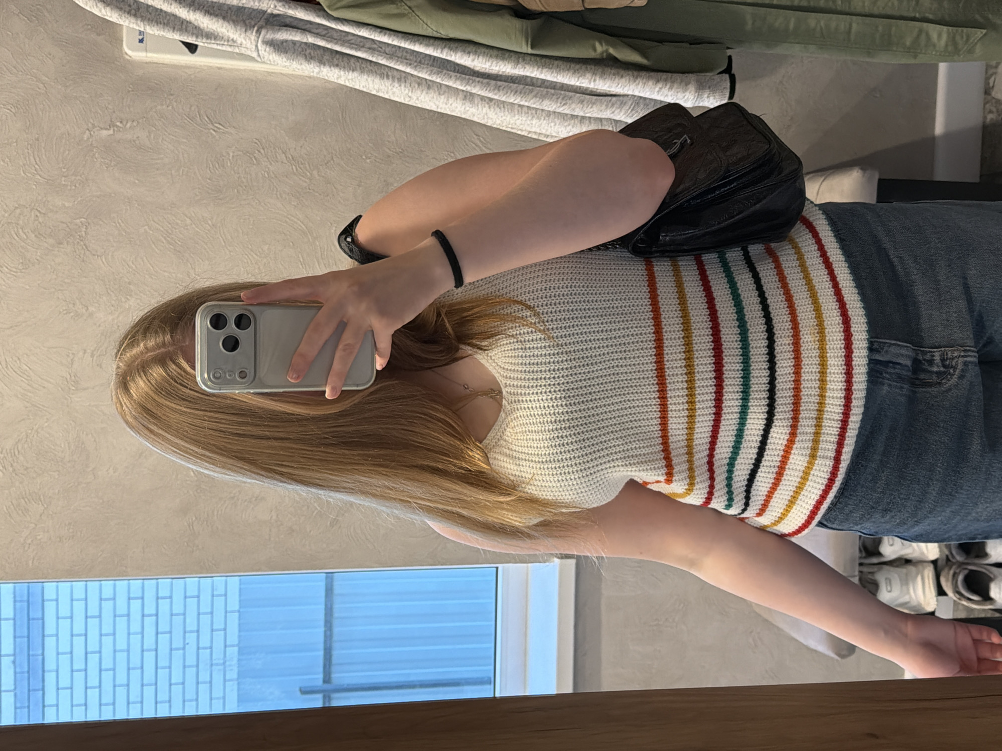 A person stands in front of a mirror, holding a smartphone to take a selfie. The person has long blonde hair and wears a white knitted top with vertical colored stripes. The person also wears blue jeans. A black backpack rests on the person's shoulder. In the background, there is a rack of clothing, a textured wall, a window, and a wooden frame around the mirror. The location appears to be indoors. The city of Rossiyskiy, Russia, cannot be determined from the image.