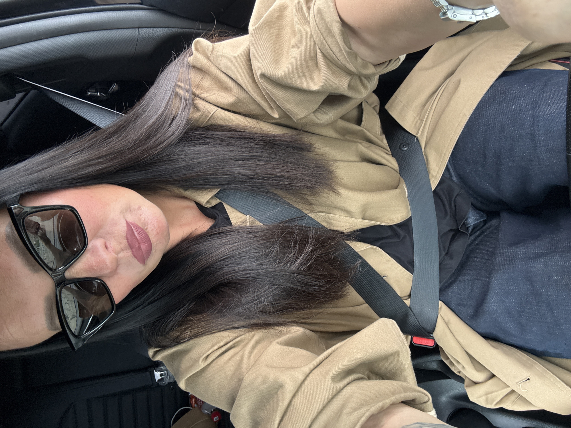 A woman is seated in the driver's seat of a car, wearing a tan jacket over a black shirt and dark blue jeans. She has on large, black, cat-eye sunglasses and her dark hair falls past her shoulders. A black seatbelt is fastened across her chest. The interior of the car is dark, with the black seatbelt mechanism and upholstery visible. The lighting suggests it is daytime, possibly overcast, with some light reflecting in her sunglasses. There is no indication of the city of Chaikovsky, Russia.