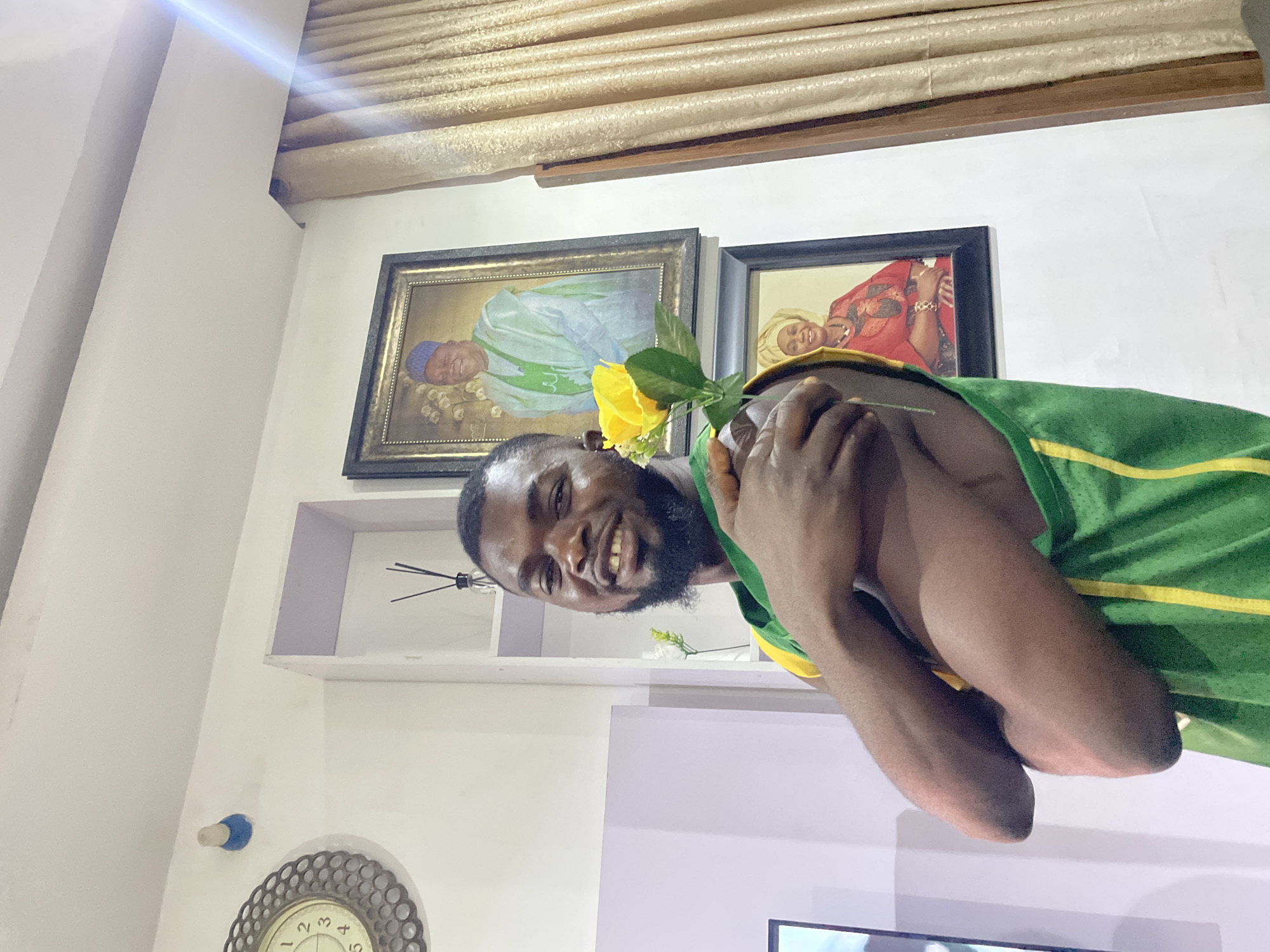 A tilted indoor portrait captures a smiling Black man in Jos, Nigeria. The man, with a dark beard and short dark hair, looks directly at the camera with a wide, joyful smile, revealing his teeth. He wears a green sleeveless top with yellow stripes and holds a single yellow artificial rose in his hands, positioned near his shoulder.
The background features white walls adorned with framed portraits. Two framed paintings hang behind him: one depicts a person in light blue and green traditional attire with a blue cap, and the other shows a person in red and gold traditional clothing, likely with a headwrap. Above these frames, a textured cream-colored curtain or blind is visible.
Below the portraits, a white shelf or alcove holds a reed diffuser. In the lower left, part of a circular wall clock with an ornate silver-colored frame and visible numbers (1 through 5) is present. A small blue and white fixture is also on the wall. A bright lens flare emanates from the upper left corner of the image, suggesting a light source. The setting appears to be a decorated home, conveying a cheerful and relaxed atmosphere.