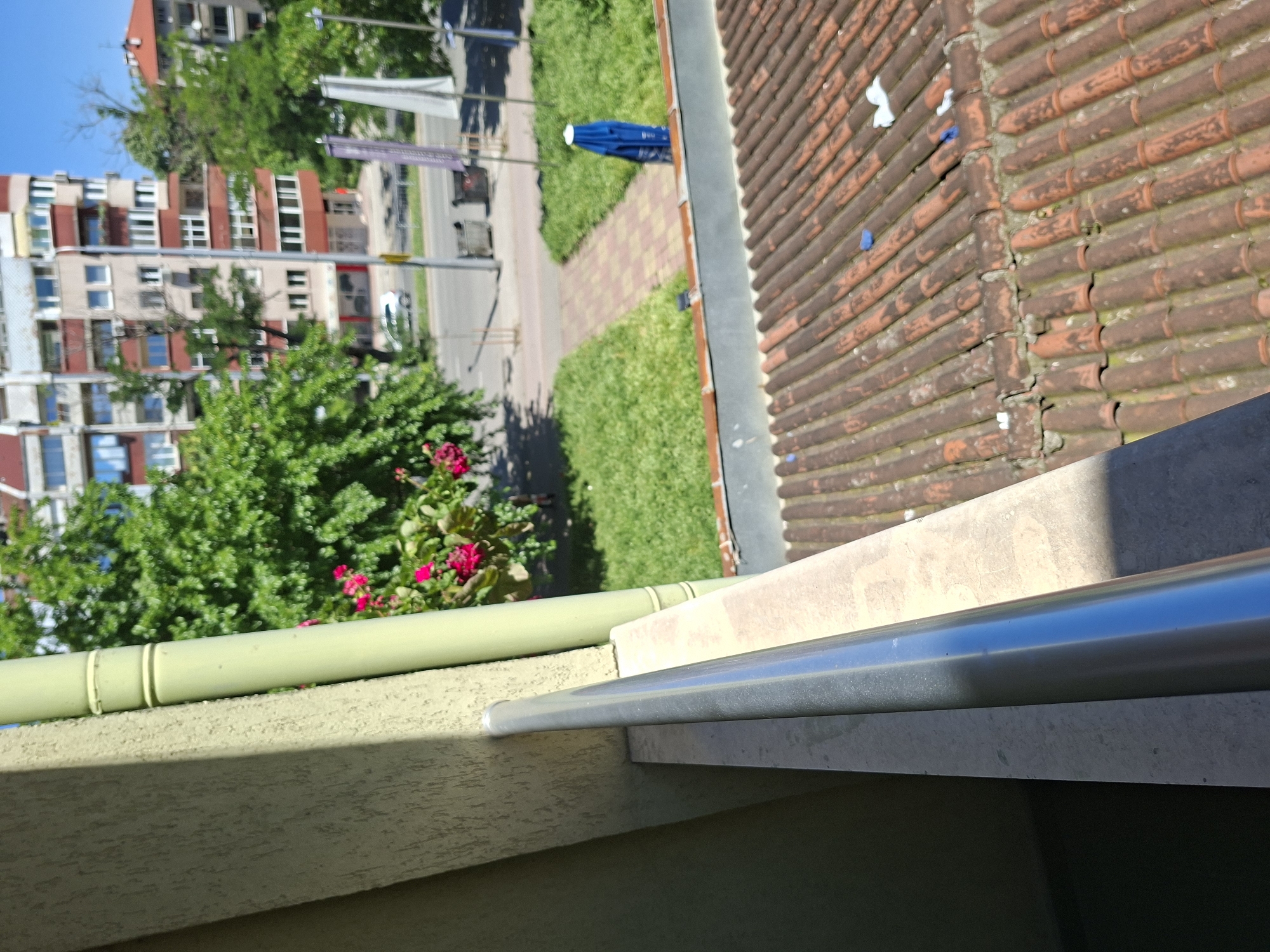 View from a balcony in Plovdiv, Bulgaria, looking outward. Buildings visible across a street lined with trees. A blue object sits on a walkway. A roof with orange tiles is to the right. A planter with pink flowers is visible on the balcony. The balcony railing is visible in the foreground.