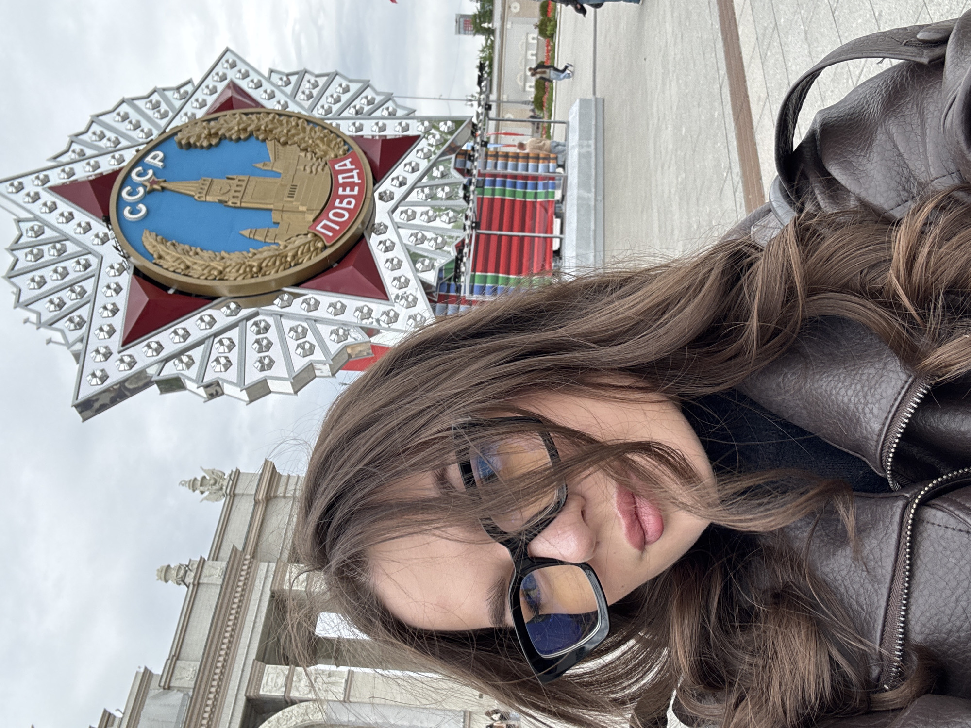 A woman with long brown hair and glasses poses for a selfie. The Victory Monument in Poklonnaya Gora, Moscow, Russia is visible in the background, featuring a large star with the Soviet emblem and the word "ПОБЕДА" (Victory) in Cyrillic script. The monument is constructed of metal and has a shiny finish. The woman is wearing a brown leather jacket and stands in front of an ornate, white building, likely part of the Victory Park complex. Other people are present in the background.