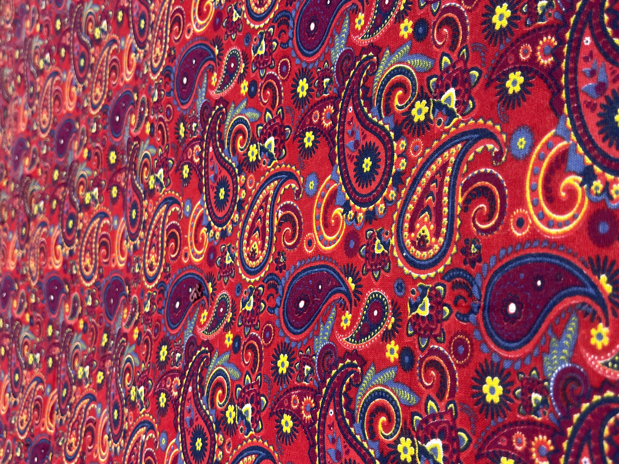 This is a close-up, low-angle, full-frame shot of a red fabric with a traditional paisley and floral pattern. The pattern features various shades of blue, yellow, and dark red, with intricate swirling designs and small flower motifs. The fabric appears to be textured, possibly cotton or a blend, and is spread out on a surface that is not visible. The lighting highlights the texture and colors of the fabric, creating a sense of depth. The image is located in Namangan City, Uzbekistan, where such textiles are commonly found.