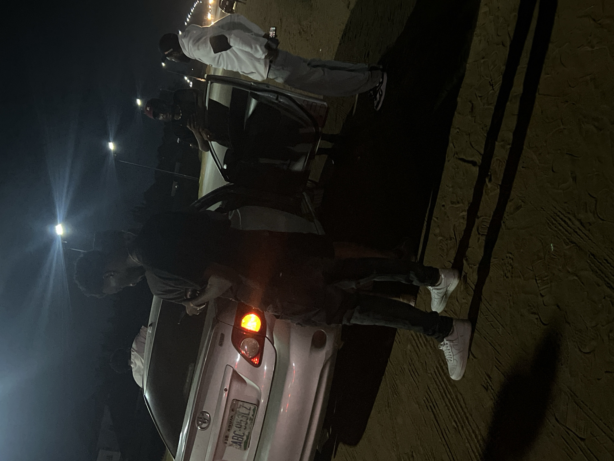 The image shows a night scene in what appears to be Galadimawa, Nigeria, based on the license plate on the back of a silver Toyota car that reads "Abuja." A young man wearing a dark shirt and pants with white sneakers is standing near the open back door of the car. There's another individual in a white outfit standing behind the car, partially obscured. Street lights illuminate the area. The ground appears sandy or uneven. The action seems focused around the car, with people potentially interacting or accessing it.