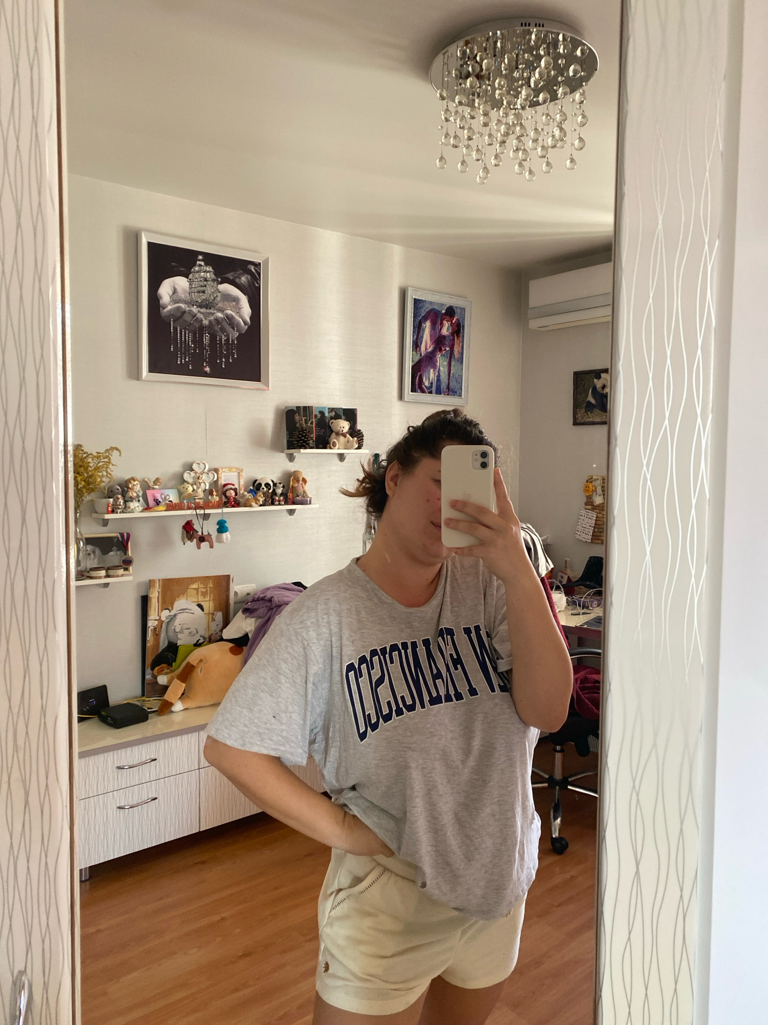 A woman stands in front of a full-length mirror in what appears to be a residential bedroom in the city of Perm, Russia. The woman is taking a selfie with her phone. She is wearing a grey t-shirt with navy blue lettering that appears to say "SAN FRANCISCO" and light-colored shorts. Behind her, reflected in the mirror, are several shelves decorated with various trinkets and figurines, a framed picture of hands holding a jeweled object, and another framed picture of a dog. A modern chandelier hangs from the ceiling. The room has wooden flooring and light-colored walls. The overall impression is a casual and personal setting within a home.