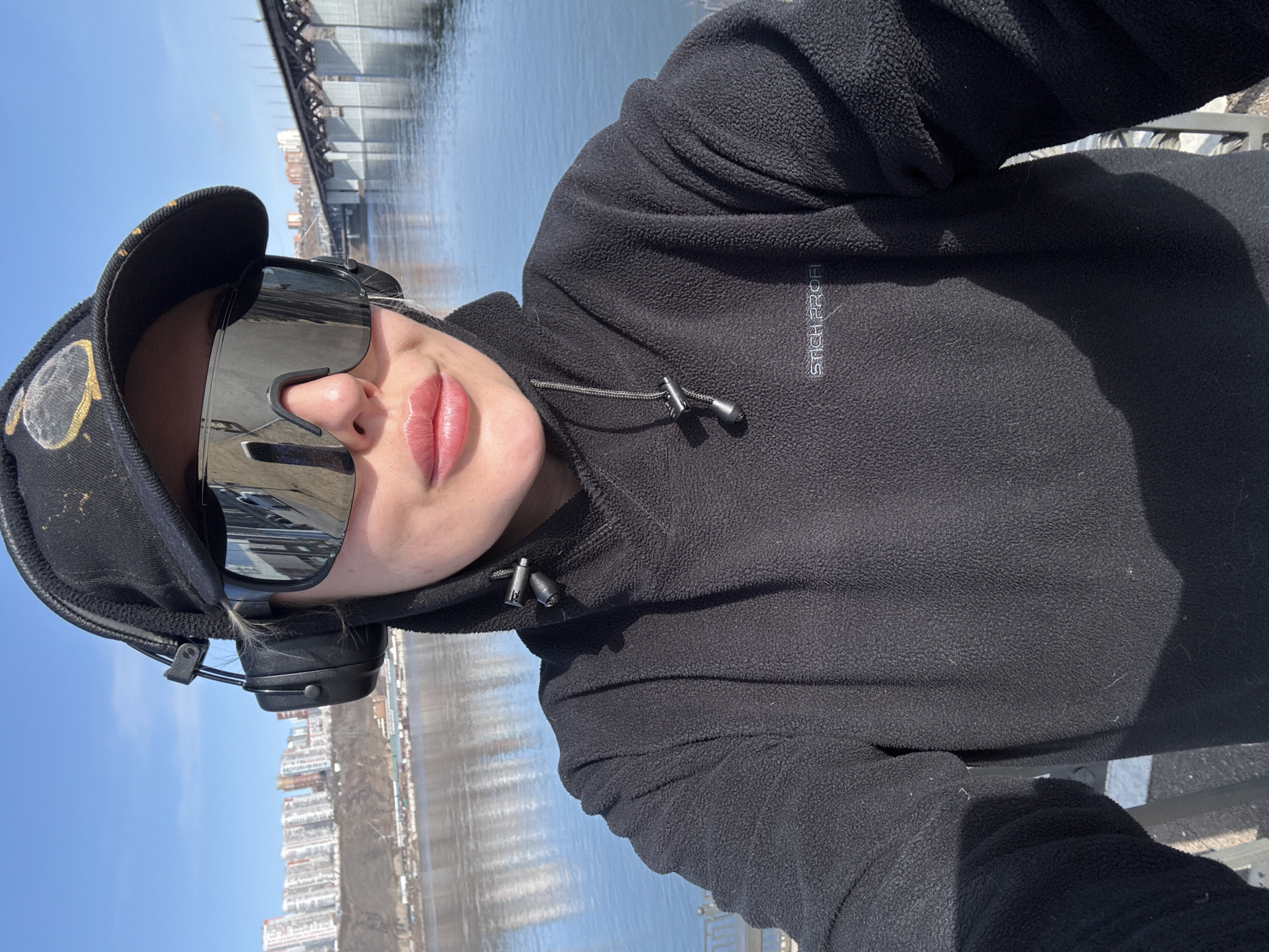 The image is a selfie of a person outdoors, likely taken on a sunny day. The person is wearing a black hooded fleece jacket, a black baseball cap, and large, mirrored sunglasses. Headphones are around their neck. The person appears to be standing by a body of water with a bridge and buildings visible in the background. The buildings and bridge design suggest that this location may be in the city of Krasnoyarsk, Russia. The water reflects the sky and surrounding buildings. The person's face is partially visible, including their nose and lips.