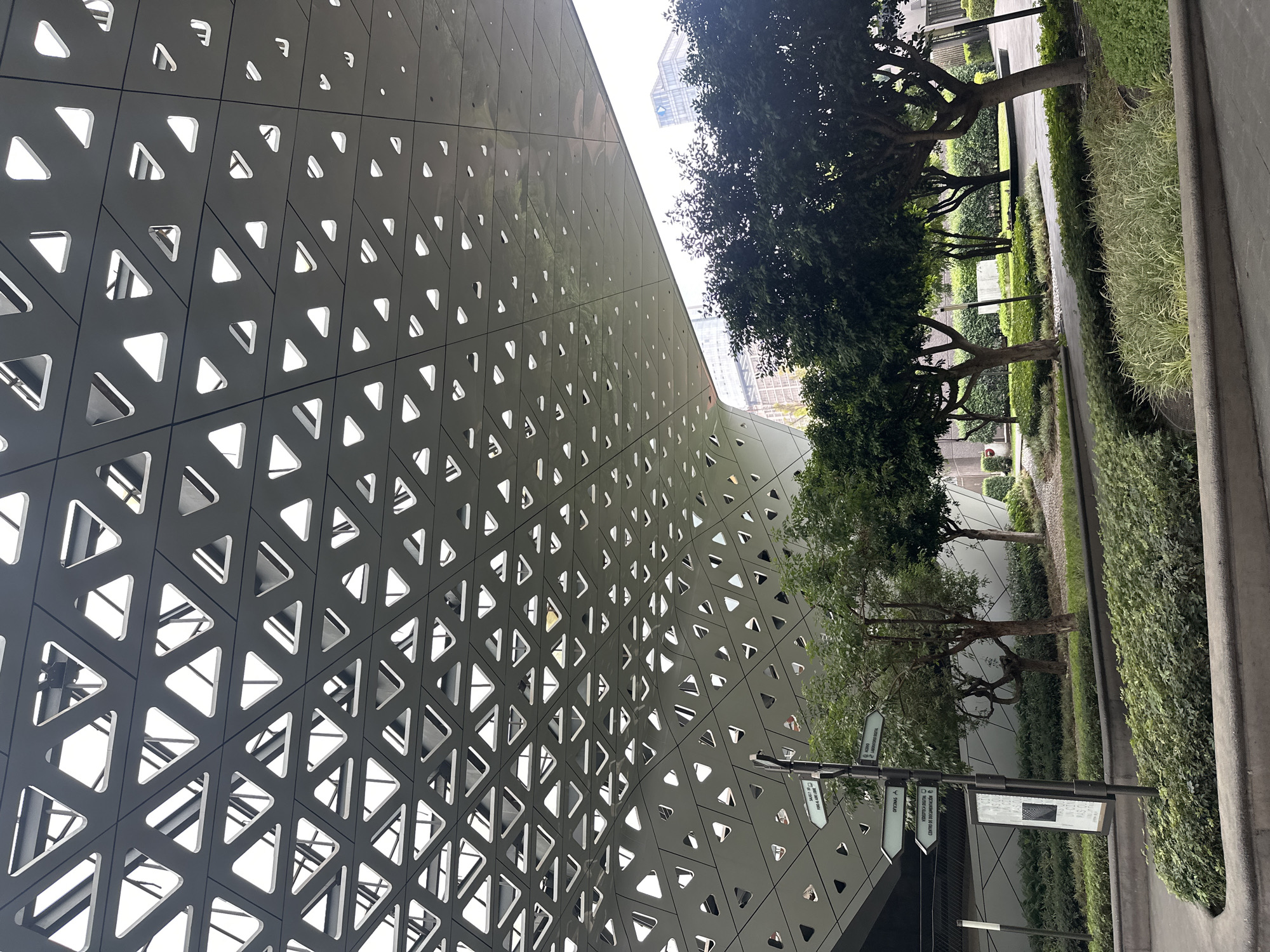 The image shows a low-angle view of the Museo Soumaya in Mexico City, Mexico. The building features a facade of interconnected, asymmetrical triangles. The color of the facade is gray. To the right of the building are trees and landscaping. In the foreground, there is a directional sign with Spanish text, planted vegetation, and a concrete walkway.
