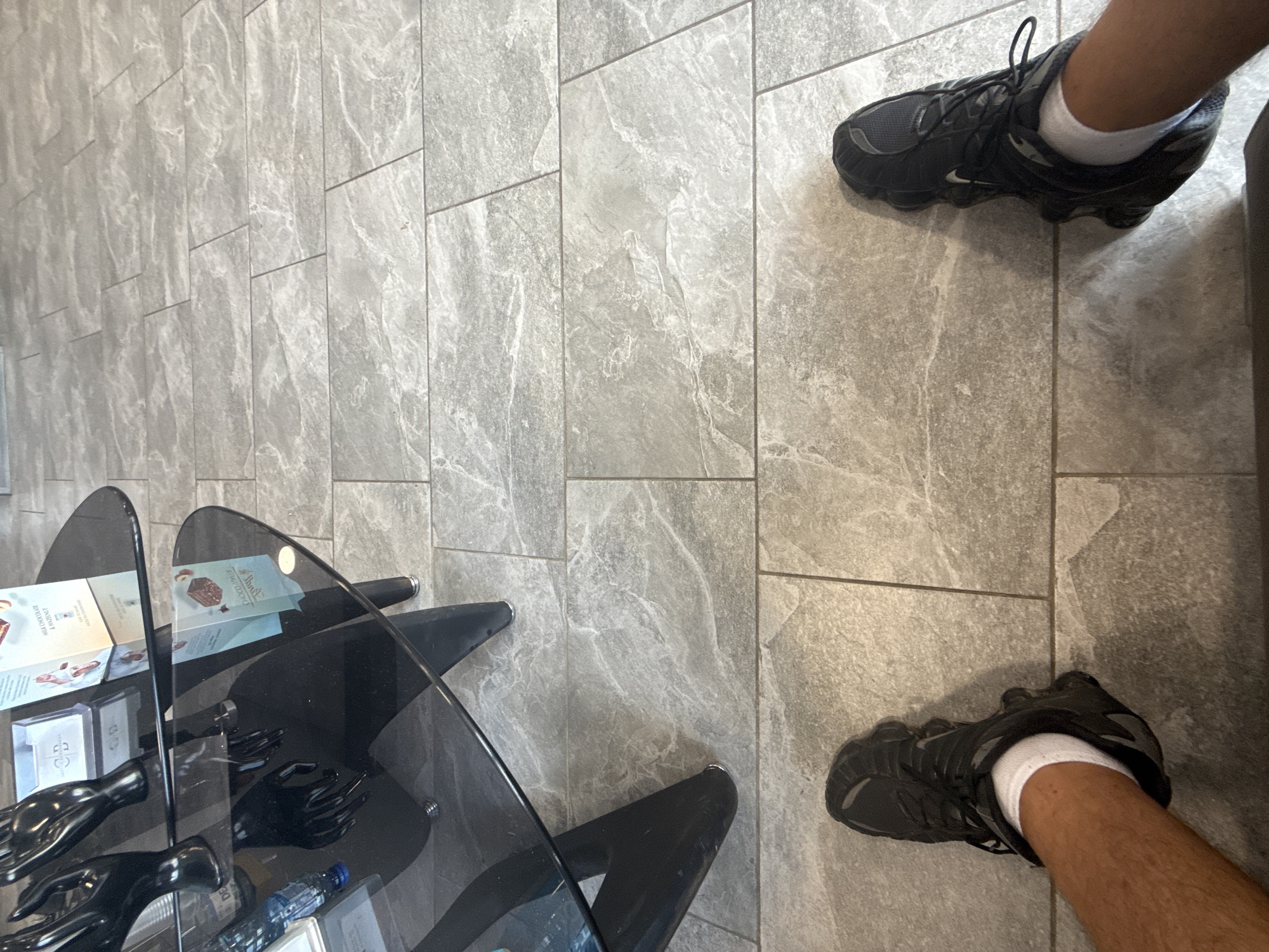 The image captures a low-angle view of a person's legs and feet wearing black athletic shoes and white socks, standing on a gray tiled floor. In the background, a modern, black, multi-tiered glass table is visible, adorned with decorative black mannequin hands and some boxes. The overall scene suggests an indoor environment, possibly a retail space or a stylish waiting area. The floor tiles have a marble-like pattern. There is no information or visual evidence to place this scene in Yambol, Bulgaria.