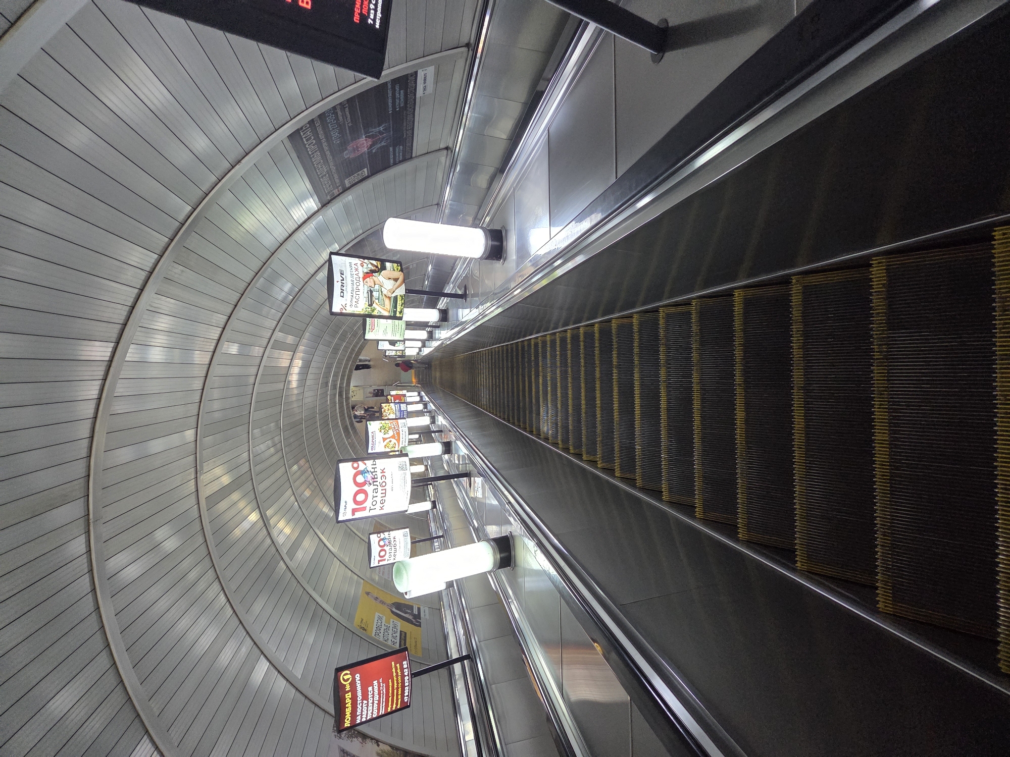 An upward-view perspective shows a wide, cavernous escalator tunnel in Yekaterinburg, Russia. The escalators, lined with stainless steel handrails and lit by tall, cylindrical white lights, descend into the distance. The arched ceiling of the tunnel is made of ribbed metal panels. Several digital billboards are mounted along the right side of the tunnel, displaying various advertisements. One billboard on the left advertises "LOMBARD NO. 1". Further down, other billboards advertise "100% total cashback" and "DRIVE Summer Sale." People are visible in the distance at the top of the escalators, descending into the tunnel. The lighting is bright and functional, highlighting the metallic surfaces and the utilitarian architecture of the space.