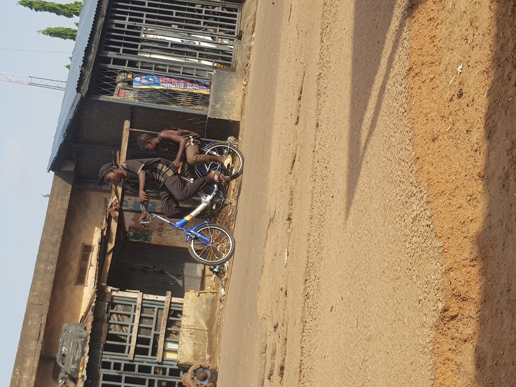 Two individuals are depicted on a bicycle on a dirt road in Kufena, Nigeria. The person in the front, wearing a black and grey striped t-shirt and black pants, is operating the bicycle. Seated behind them, facing forward, is another individual wearing a dark sleeveless shirt and light brown pants. The bicycle has a blue frame and white tires with yellow rims. In the background, a building with a corrugated metal roof and bars on its windows is visible. The environment appears to be an outdoor, possibly urban or semi-urban setting, characterized by the unpaved road and the utilitarian nature of the building.