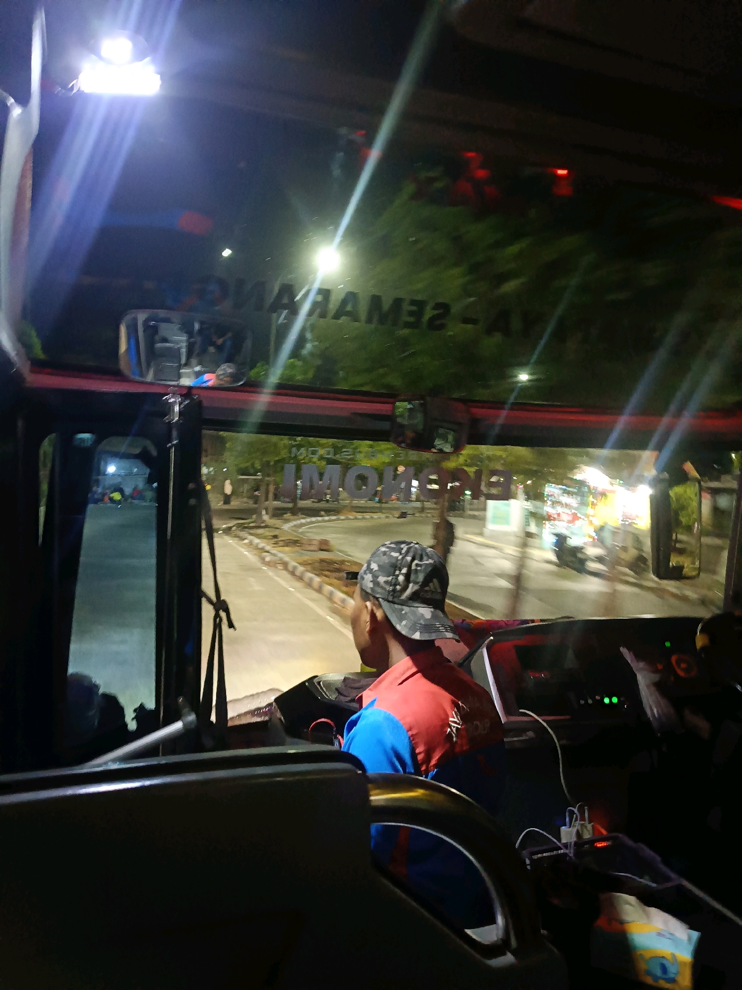 A man wearing a camouflage baseball cap and a red and blue uniform sits in the driver's seat of a bus at night. The bus appears to be at a terminal or station, possibly in Bungurasih, Indonesia, given the signage reflected in the windshield. The reflection shows what appears to be text that reads "MAYA-SEMARANG". The driver's hands are not visible, but the steering wheel and dashboard are in front of him. The interior of the bus is dimly lit with some overhead lights and lights from the dashboard. Outside the bus, streetlights and building lights are visible, along with trees and other vehicles. Another person is visible in the reflection on the left side of the windshield, and there are other people and activity visible through the side window. A charging cable and adapter are plugged into a device on the dashboard.