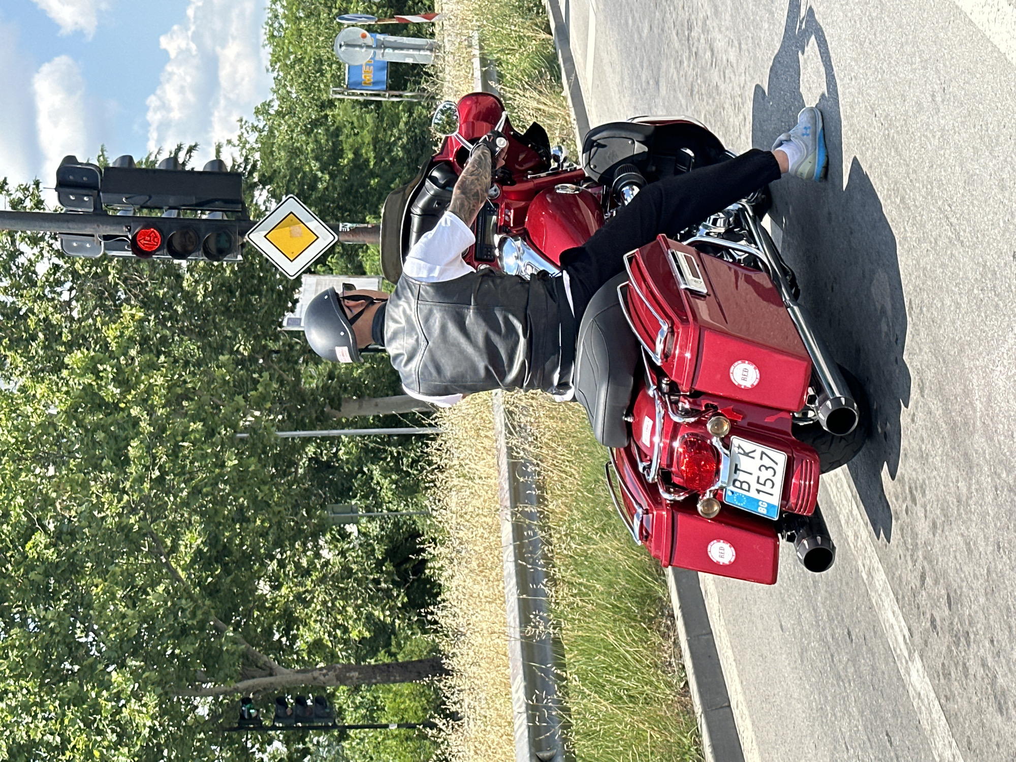 The image shows a person riding a red motorcycle. The rider is wearing a black leather vest over a white shirt, black pants, a black helmet, and sunglasses. The motorcycle is a touring bike with saddlebags. The rider has tattoos on their arm.
The motorcycle is on a paved road, with grass and trees on the side. A traffic light is visible in the background, and it is red. A diamond-shaped road sign with a yellow diamond is also present. The time of day appears to be daytime, with bright sunlight casting shadows. The weather seems clear and sunny.
The license plate on the back of the motorcycle reads "BT K 1537" and has a European Union symbol. There are also round red stickers on the motorcycle that say "RED". The scene is likely a street in Veliko Tarnovo, Bulgaria. The rider appears to be waiting at a traffic light, possibly contemplating their next move.