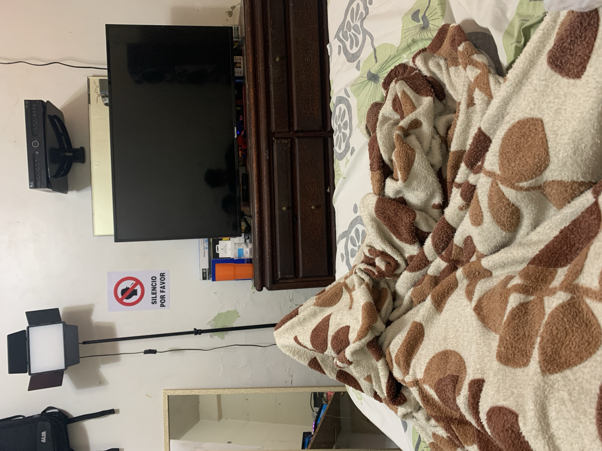 The image depicts an interior scene, possibly a bedroom, with a television mounted on the wall to the left. Adjacent to the television is a dark brown wooden dresser. Below the TV and slightly to the left is a light panel on a stand, situated above a framed mirror on the lower left. A sign reading "Silencio Por Favor" (Silence Please) is affixed to the wall between the TV and the light panel. To the right, covering a significant portion of the image, is a bed with a light-colored patterned bedsheet and a plush blanket with brown leaf-like patterns. A black bag is visible on the lower left corner. The location, El Valle del Espíritu Santo, Venezuela, cannot be confirmed from the image alone. No people are visible in the image.