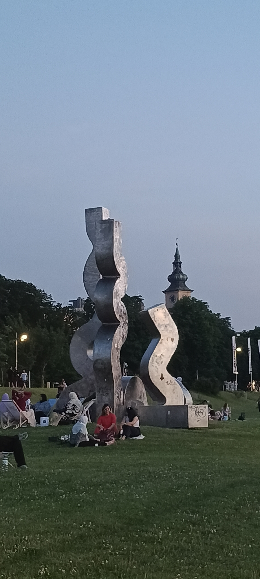 In the city of Linz, Austria, a public park scene unfolds against a dusky sky, dominated by a large, abstract metal sculpture with undulating, wave-like forms. Several individuals are scattered across the grassy terrain, engaged in various activities. In the foreground, a group of people sits on blankets and in low chairs, appearing to relax and socialize. One person in a red top and patterned skirt sits cross-legged, facing away from the viewer. To their right, another individual, wearing a white top and dark skirt, is seated. In the middle ground, a person in a red shirt is also seated on the grass. Further back, more people are visible, some seated and others standing, interspersed with the trees that form a dense backdrop. The sculpture itself is the primary focal point, its metallic surface reflecting the ambient light. Behind the sculpture and trees, the spire of a clock tower is visible, suggesting the presence of a historical building within the urban landscape. The overall atmosphere is one of leisure and public gathering in an outdoor setting.
