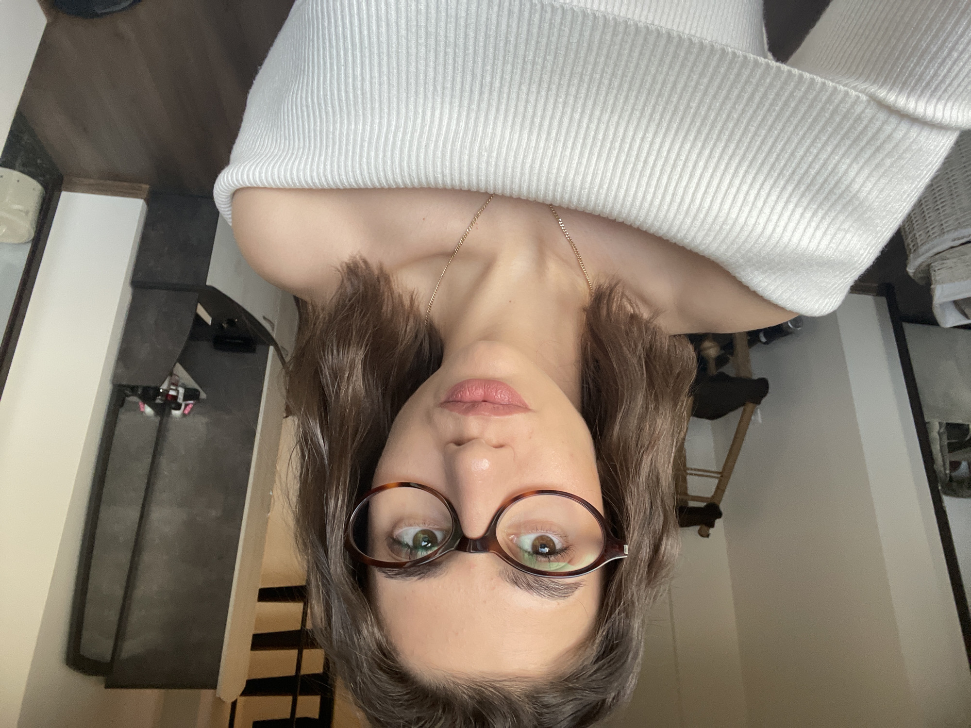The image is an upside-down, close-up selfie of a woman wearing brown-rimmed glasses and a white, off-the-shoulder sweater. A gold chain necklace is visible around her neck. She has light skin, brown hair, and is looking directly at the camera.
The background is blurry, but elements of an interior space can be discerned. There's a stairway with dark treads, a mirror on a wall, and a wooden chair. Given the interior elements, the location is likely inside a residential building. There's not enough visual information to determine if the location is Sofia, Bulgaria.