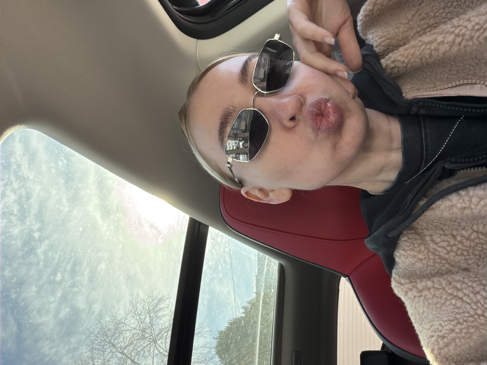 The image displays a selfie taken inside a vehicle, presumed to be in Chaikovsky, Russia, although no specific landmarks are identifiable. A blonde-haired individual with light skin is centrally positioned, wearing silver-framed, hexagonal sunglasses. Their lips are pursed in a pouting expression, and their left hand rests on their chin. They are dressed in a black hooded sweatshirt beneath a light brown, textured, sherpa-style jacket, accessorized with a thin silver chain necklace. The vehicle's interior features a red and black sport-style seat and a beige headliner/A-pillar. Through the car's panoramic sunroof, a partly cloudy sky with bright sunlight and a noticeable lens flare is visible, along with the silhouette of a bare-branched tree. The side window reveals a different tree with denser foliage and a distant, indistinct structure. Reflections within the sunglasses depict a street environment including parked cars, tree trunks, and urban buildings.