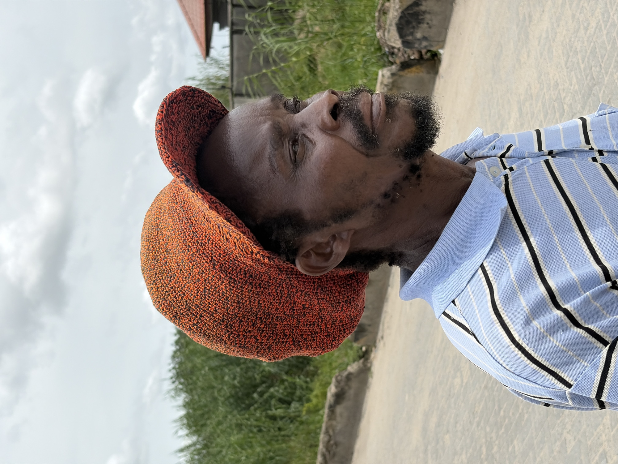 The image presents a close-up, side profile shot of an adult Black man looking upwards. He is wearing a distinctive orange, textured, knitted cap and a light blue polo shirt adorned with vertical black and thin white stripes. The man has a full beard and mustache. His eyes are open, and his gaze is directed towards the upper right of the frame (when upright). The background is an outdoor scene under a cloudy sky, suggesting daytime. Green foliage, likely bushes or trees, is visible behind him to his left, and a paved or concrete surface stretches into the background. A hint of a building or structure can be seen in the distance on the upper right. The man's expression is neutral. No visible text is present. The setting is Igboefon, Nigeria.