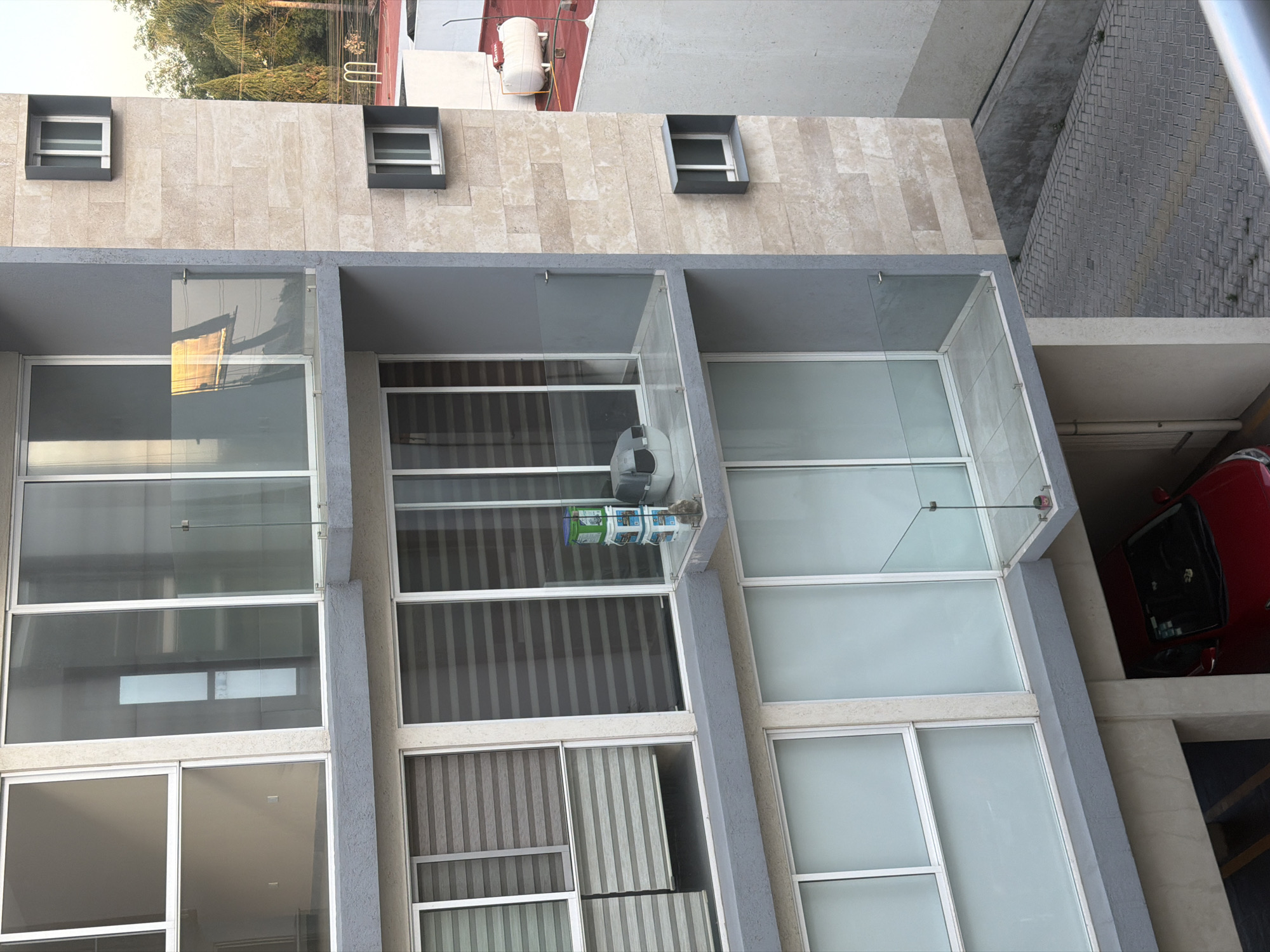 An exterior view captures a modern, multi-story building, likely an apartment or office complex, from an elevated perspective looking slightly downwards. The building features a light-colored, stone-tiled upper facade with two small, rectangular, dark-framed windows set into pronounced boxes.
Below this, several levels are characterized by large rectangular windows with white frames and recessed balcony-like areas, many fitted with glass railings. The primary structural elements of these lower levels are composed of grey concrete or stucco. Some windows reveal internal blinds or shades with horizontal stripes. In one of the recessed areas on a middle floor, a small, colorful object, possibly a plant holder made of stacked containers, hangs from the ceiling. Reflections of sunlight, green trees, and potentially other structures are visible in the glass surfaces of the windows and railings.
At the ground level, to the right of the building, there is a section of gray, cobblestone-like paving. Below the building itself, a red car is partially visible, parked in what appears to be an underground or covered parking area. In the background, above the tiled facade, lush green trees and parts of other buildings or roofs are discernible, including a white, cylindrical water heater mounted on a red roof surface.
The scene is brightly illuminated, indicating daytime. No people or active events are visible, presenting a static architectural study of the building and its immediate surroundings. There is no visible text. The image provides a detailed look at the building's facade and adjacent areas, characteristic of urban environments in places like San Andrés Cholula, Mexico.