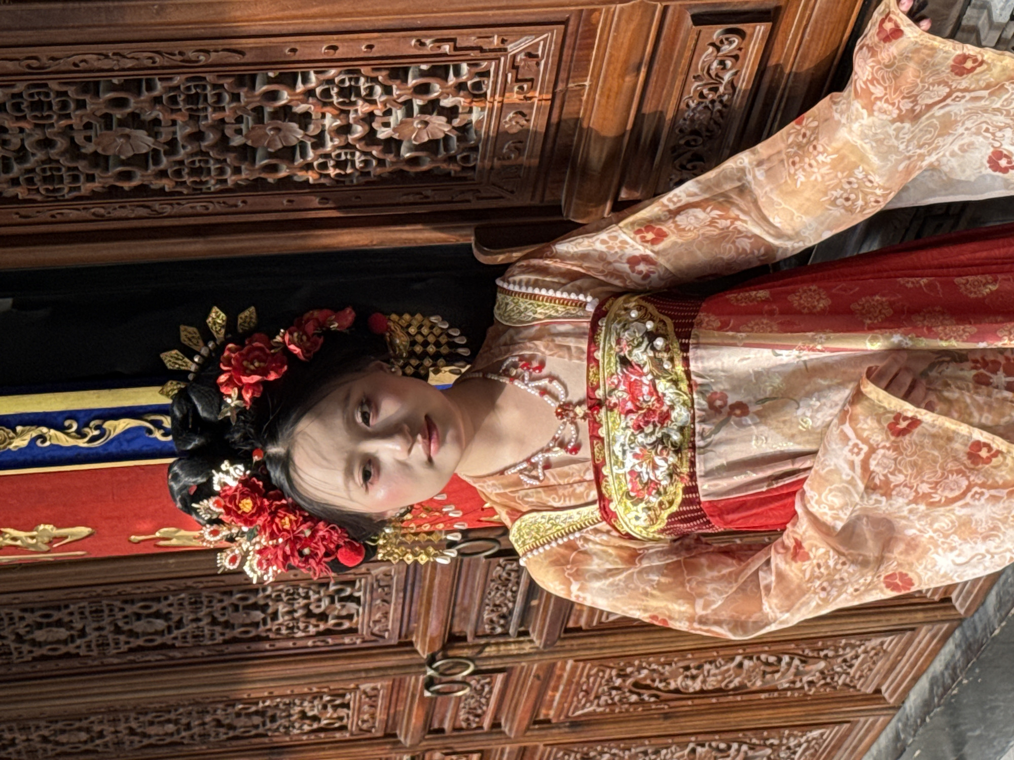 A person wearing a traditional Chinese hanfu dress poses next to an elaborately carved wooden cabinet or wall. The person's dark hair is styled with red floral ornaments and gold-colored accents. The hanfu features intricate patterns and embroidery in colors of red, gold, and cream. The background appears to be an interior space with traditional Chinese furniture and decorations. It is likely located in Yuzhong District, China, based on the context of the image.