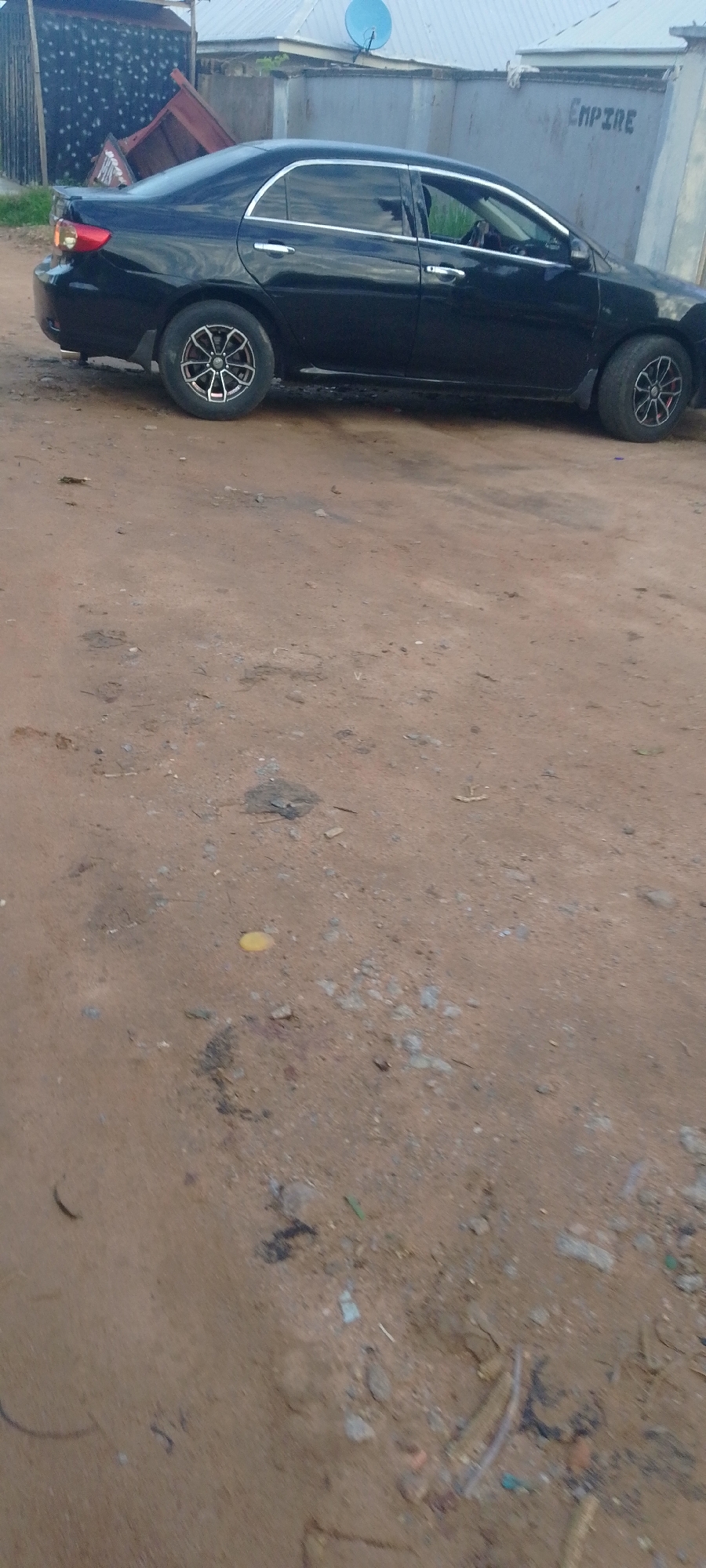 A black sedan is parked on a dirt road in Jalingo, Nigeria. The car is a Toyota Corolla, model year 2008-2013, with chrome accents and alloy wheels. The car's right side is visible, facing left. The background shows a building with a gray corrugated metal roof and a blue satellite dish. To the left of the car, a red truck bed is visible, leaning against a dark, textured wall. The ground is sandy with scattered debris, including small rocks, twigs, and some dark stains. The sky is not visible in the frame. The image appears to have been taken during daylight.