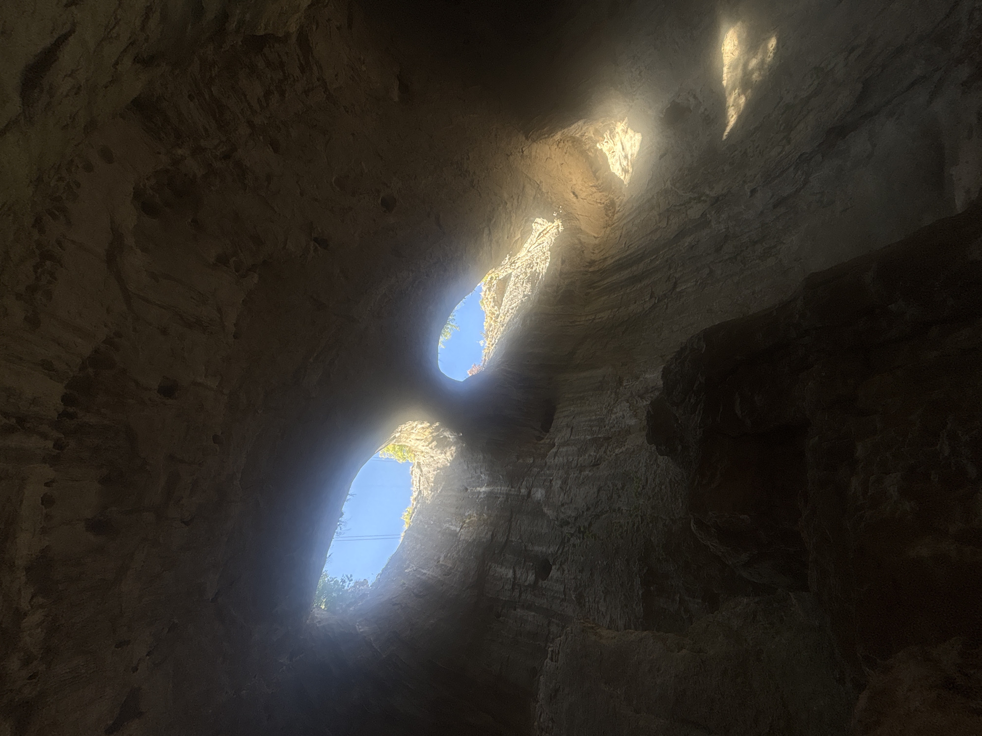The image captures the Prohodna Cave, also known as the Eyes of God cave, located in Karlukovo, Bulgaria. The cave features two large, eye-shaped openings in its ceiling, through which sunlight streams into the cavern. The rock formations within the cave are rough and textured, with visible striations indicating geological processes. Vegetation can be seen growing around the edges of the openings in the ceiling. The overall impression is one of natural grandeur and a unique geological formation.