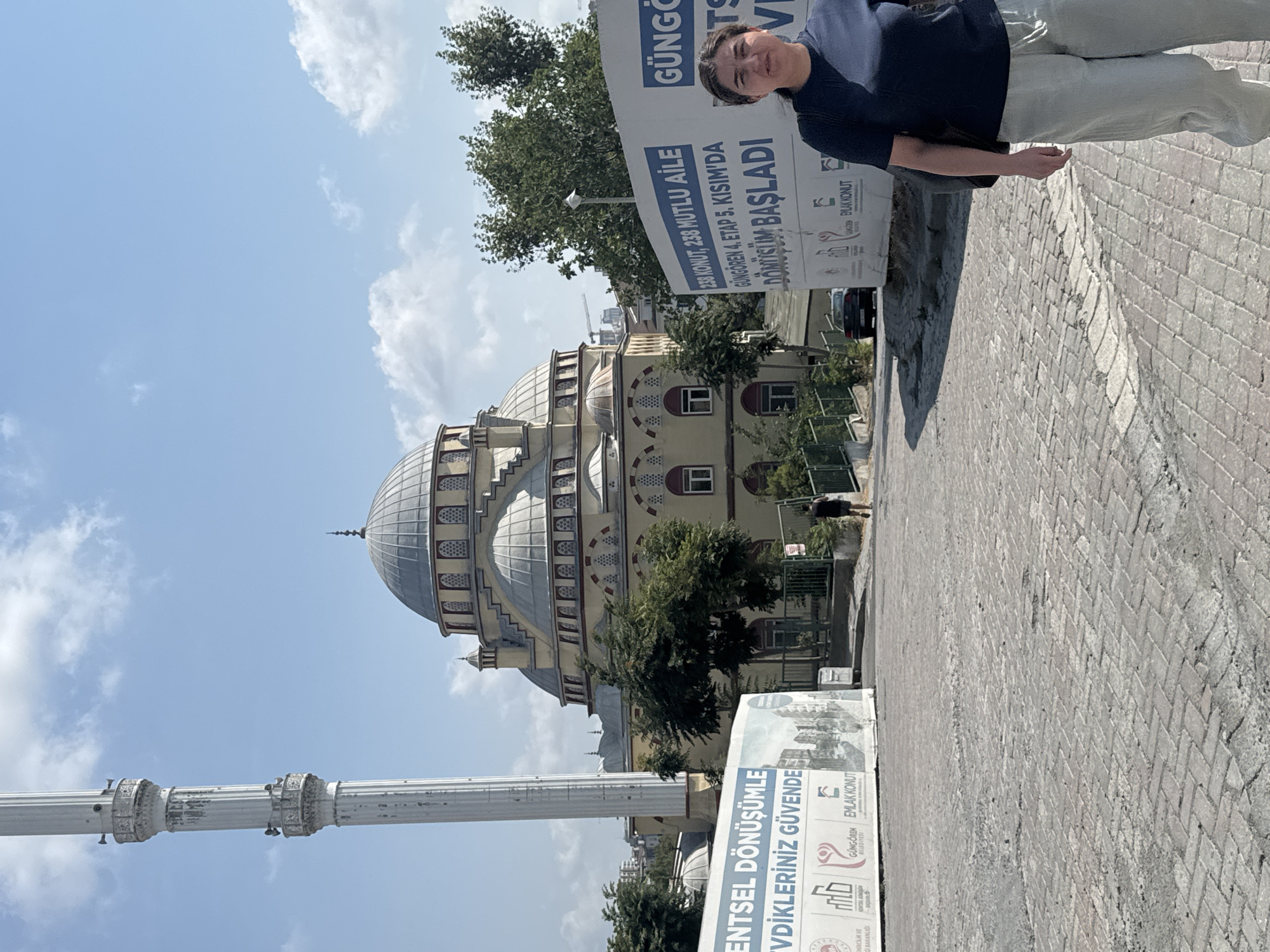 The image shows a woman standing on a cobblestone street in front of a mosque and a construction banner. The mosque has a large dome and a tall minaret. The banner advertises a residential development called "Dönüşüm Başladı" (Transformation Has Begun) in Güngören, Turkey, with text mentioning "238 KONUT, 238 MUTLU AİLE" (238 UNITS, 238 HAPPY FAMILIES) and "GÜNGÖREN 4. ETAP 5. KISIM'DA" (In Güngören, Stage 4, Phase 5). The woman is wearing a dark blue t-shirt and light-colored pants, and is carrying a black bag. In the background, there are trees, a car, and a cloudy blue sky. A man is walking away from the camera in the middle distance.