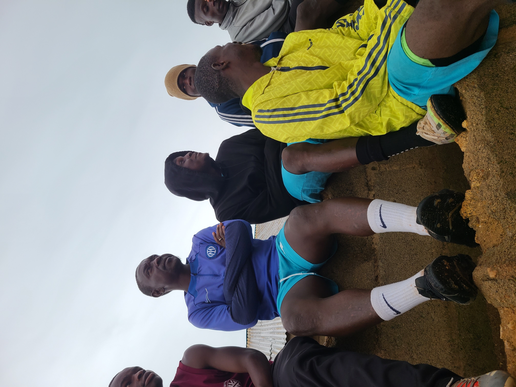 The image, rotated 90 degrees clockwise, captures a low-angle perspective of at least six men resting outdoors against a light, overcast sky in Rafin Bauna, Nigeria. The men are situated on rugged, sandy, and earthy terrain, looking upwards.
In the mid-ground, a man wears a vibrant yellow Adidas jacket with distinctive blue stripes on the sleeves and blue athletic shorts. His light-colored athletic shoes are partially visible against the ground. To his left, another man is seen in a black hooded sweatshirt and blue shorts. Behind them, a man in a grey hooded top with blue stripes and another wearing a brown beanie are also looking up.
In the foreground, on the left side, a man in a blue long-sleeved quarter-zip top, adorned with the Inter Milan football club logo on the chest, has his arms crossed while looking upwards. He is wearing blue athletic shorts, white socks featuring a black Nike swoosh, and dark athletic shoes. Another man, partially visible in the bottom left, is wearing a maroon t-shirt.
The scene suggests a moment of rest or observation, possibly during a sports event or training session, given the athletic attire. The sky is uniformly bright grey, indicating an overcast day. The ground texture is coarse, characteristic of natural, unpaved outdoor areas. No strong emotions are overtly displayed, with expressions generally neutral as they gaze skyward.