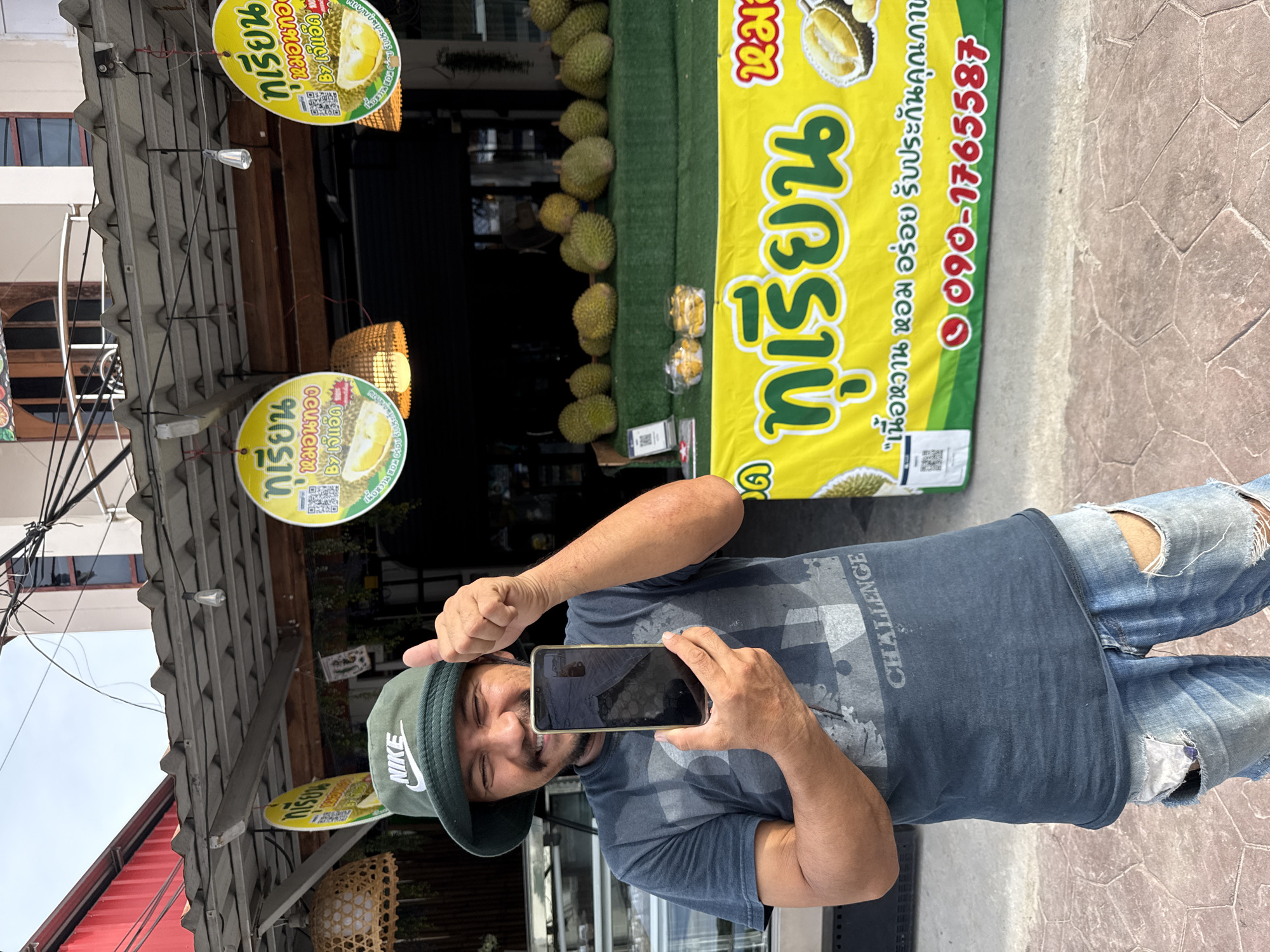 A man stands outdoors, holding a smartphone, in front of a durian stand in Moo 1, Thailand. He is wearing a green Nike hat, a grey-blue t-shirt with "CHALLENGE" printed on it, and ripped blue jeans. The durian stand is displaying numerous durians, along with yellow signs featuring Thai script and images of durians. One sign includes a phone number, 090-1765587. Hanging above are round signs with similar imagery. The environment appears to be a commercial area.