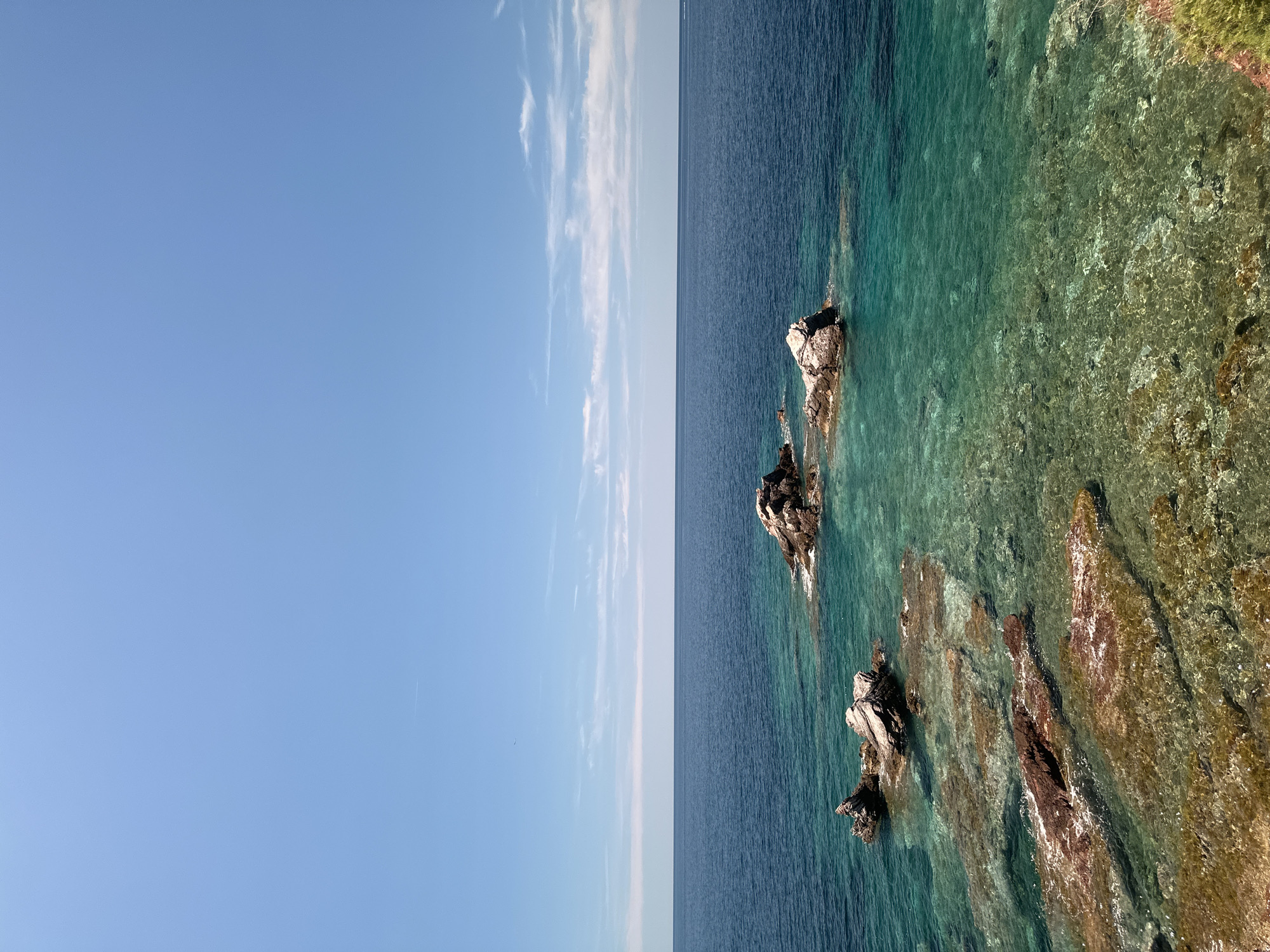 The image depicts a serene coastal scene in Sveti Stefan, Montenegro. The foreground is dominated by shallow, crystal-clear turquoise water revealing submerged rocks and a sandy bottom. Several larger, weathered rocks protrude from the water closer to the shore. Further out, a cluster of three rocks rises from the deeper, dark blue sea. The horizon line is clearly defined, separating the vast expanse of the sea from a bright blue sky dotted with wispy white clouds. A faint contrail from an aircraft is visible in the upper portion of the sky. The overall impression is one of natural beauty and tranquility, characteristic of the Adriatic coast.