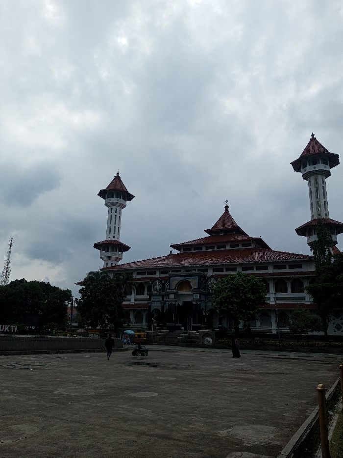 The image displays a large mosque situated in the city of Daringbadi, India, beneath an overcast sky. The white-walled mosque features a multi-tiered red-tiled roof system and is flanked by two tall, slender minarets. Each minaret possesses multiple open balconies and is capped with a red conical roof. A dark, ornate entrance is centered on the building's facade. In the foreground, a grey, open plaza extends, partially obscured by shadows. One individual, dressed in dark clothing, walks across the plaza, moving away from the viewer towards the mosque. A second individual stands further right, closer to the mosque's base. A small, colorful wheeled cart or ride with a blue umbrella is located in the center-left of the plaza. Dense green trees and shrubs line the mosque's perimeter and the plaza's edges. On the far left, a tall, dark telecommunications lattice tower and a partial sign reading 'BUKTI' are visible behind foliage.