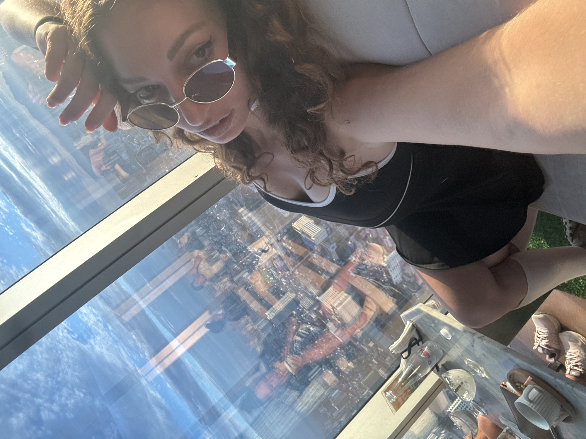 The image shows a woman taking a selfie in a high-rise building overlooking a city. The woman is wearing sunglasses and a black dress with white trim. She has curly brown hair and her hand is raised to her forehead. In the background, the city skyline is visible through a large window. The reflection in the window shows other people and the interior of the room. There is a table with drinks and napkins on it. The image appears to be taken during the daytime, with sunlight streaming in through the window. The specific location in Hanoi, Vietnam cannot be determined from the image alone.