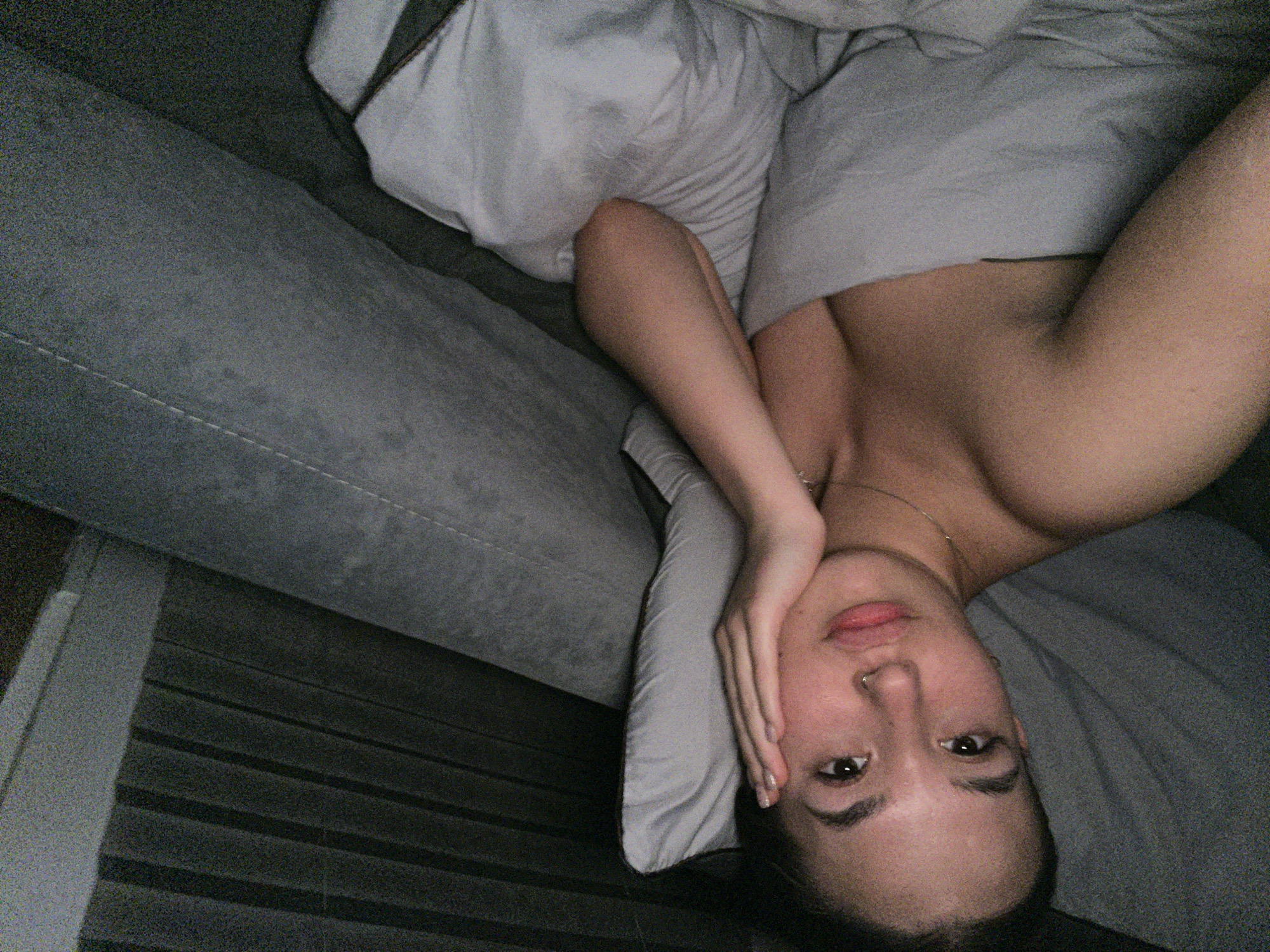 A woman is lying on a bed or couch covered in light gray bedding. Her head is on a pillow, and she is turned sideways with her face facing the camera. Her right hand is resting on her cheek. She is wearing a necklace. A gray sofa is visible on the left. The image appears to have been taken indoors, likely in a private residence. There is no evidence to confirm that the image was taken in Lobanovo, Russia.