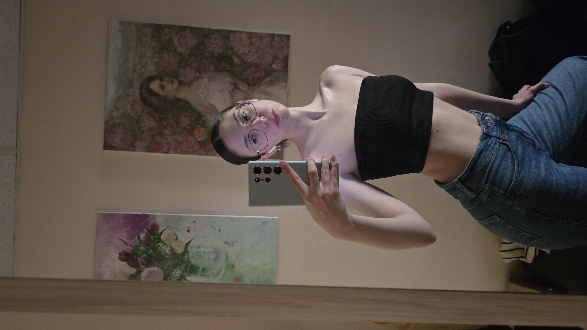 A young woman takes a selfie in front of a mirror. She is wearing a black tube top and blue jeans. She is holding a light blue or silver smartphone, possibly a Samsung Galaxy S22 Ultra, in her right hand. The woman has short dark hair, glasses, and is wearing small earrings.
Behind her, on the wall, are two framed art pieces. The top one depicts a reclining woman amidst a floral setting, likely roses. The bottom artwork shows a vase of flowers with shades of purple and green.
The overall environment appears to be an interior space, possibly a bedroom or hallway, with light-colored walls. There is a wooden frame surrounding the mirror. Part of a dark-colored bag is also visible on the right side of the image.
There is no indication of Stara Zagora, Bulgaria.