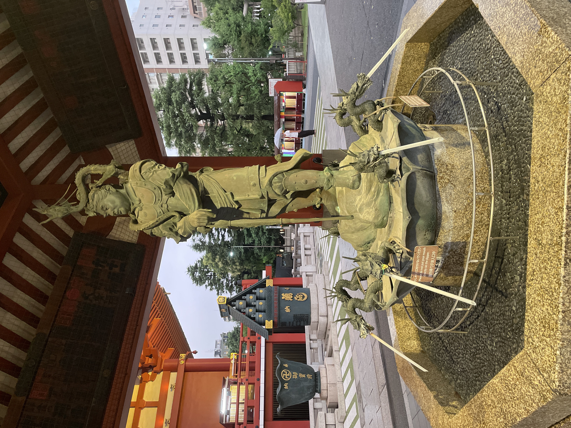 In Taito, Japan, a large bronze statue of a warrior stands prominently in the foreground, positioned within the covered entryway of a traditional Japanese temple or shrine. The statue depicts a figure in ornate armor, with a dragon coiled around its head and shoulders, holding a staff planted in the ground. In front of the warrior statue, a water basin features several dragon spouts, from which streams of water are flowing into the basin filled with dark pebbles. The basin itself is surrounded by a golden-hued, octagonal stone structure. Behind the water basin, to the left, is a large, dark bronze pot with intricate carvings, and to the right, a dark green, pagoda-like structure with golden trim, adorned with what appear to be stacked cups. The background reveals a bustling city scene with trees, a modern multi-story building, and a bright red kiosk-like structure where a person holding a white umbrella is visible. The sky is overcast, suggesting either early morning or late evening. The overall scene blends ancient religious iconography with the modern urban environment.