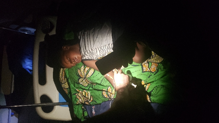 The scene depicts a medical intervention in Narayi, Nigeria, occurring in low light conditions. A young, dark-skinned child, approximately 5-10 years old, lies on a medical bed, covered by a green patterned blanket, appearing to be asleep or unconscious. The child wears a grey and white striped tank top. An intravenous line is inserted into the child's left forearm, secured with white tape. A light-skinned adult hand on the left is holding a syringe, connected to this IV line, actively administering a substance. Another hand, dark-skinned, is visible on the right, holding a black smartphone, with its screen illuminated, potentially providing additional light to the immediate area. A white hospital bed frame and an IV pole are partially visible in the dimly lit background.