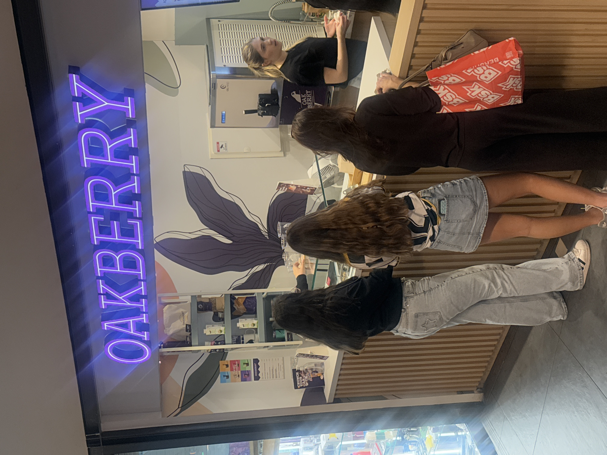 Three female patrons stand at the counter of an "Oakberry" store. A female employee is behind the counter. The storefront is identified by a vertically-oriented, neon purple "Oakberry" sign. Inside, there is shelving with products for sale and a decorative wall mural of a dark purple flower. One patron has a red "BSK" bag. This scene is in the city of Matosinhos, Portugal.