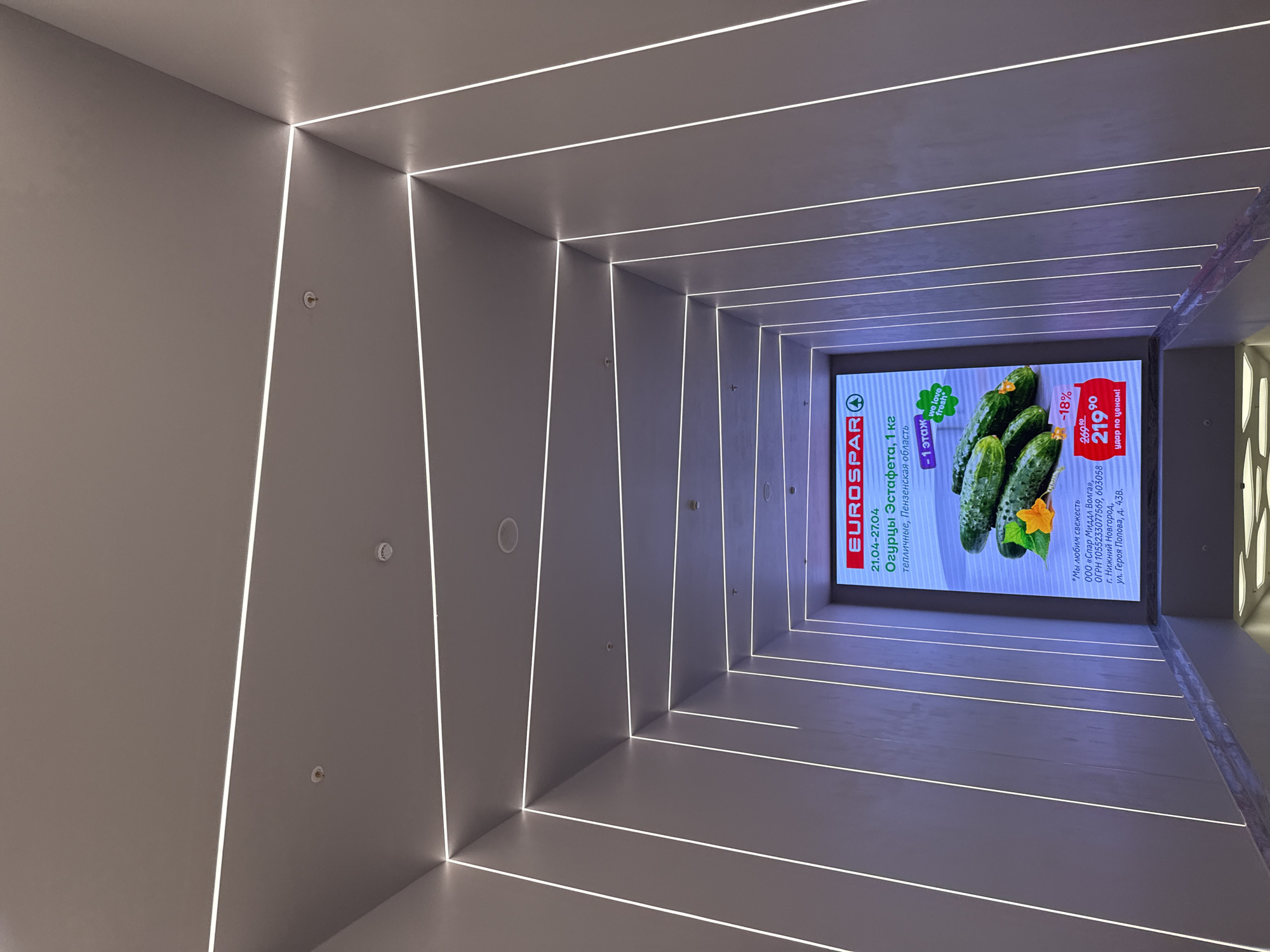 This is a low-angle indoor shot of a modern corridor with recessed lighting on the walls and ceiling, leading to a Eurospar advertisement at the end. The corridor walls are a neutral tone, with recessed linear lighting extending inward. The advertisement at the end features cucumbers and information about a promotion. Text is visible, including references to Eurospar, the dates 21.04-27.04, "Огурцы Эстафета, 1 кг", a price of 219.90, and mentions of Penza Oblast and Nizhny Novgorod. The precise location of this scene in Moscow, Russia cannot be determined from the image alone.