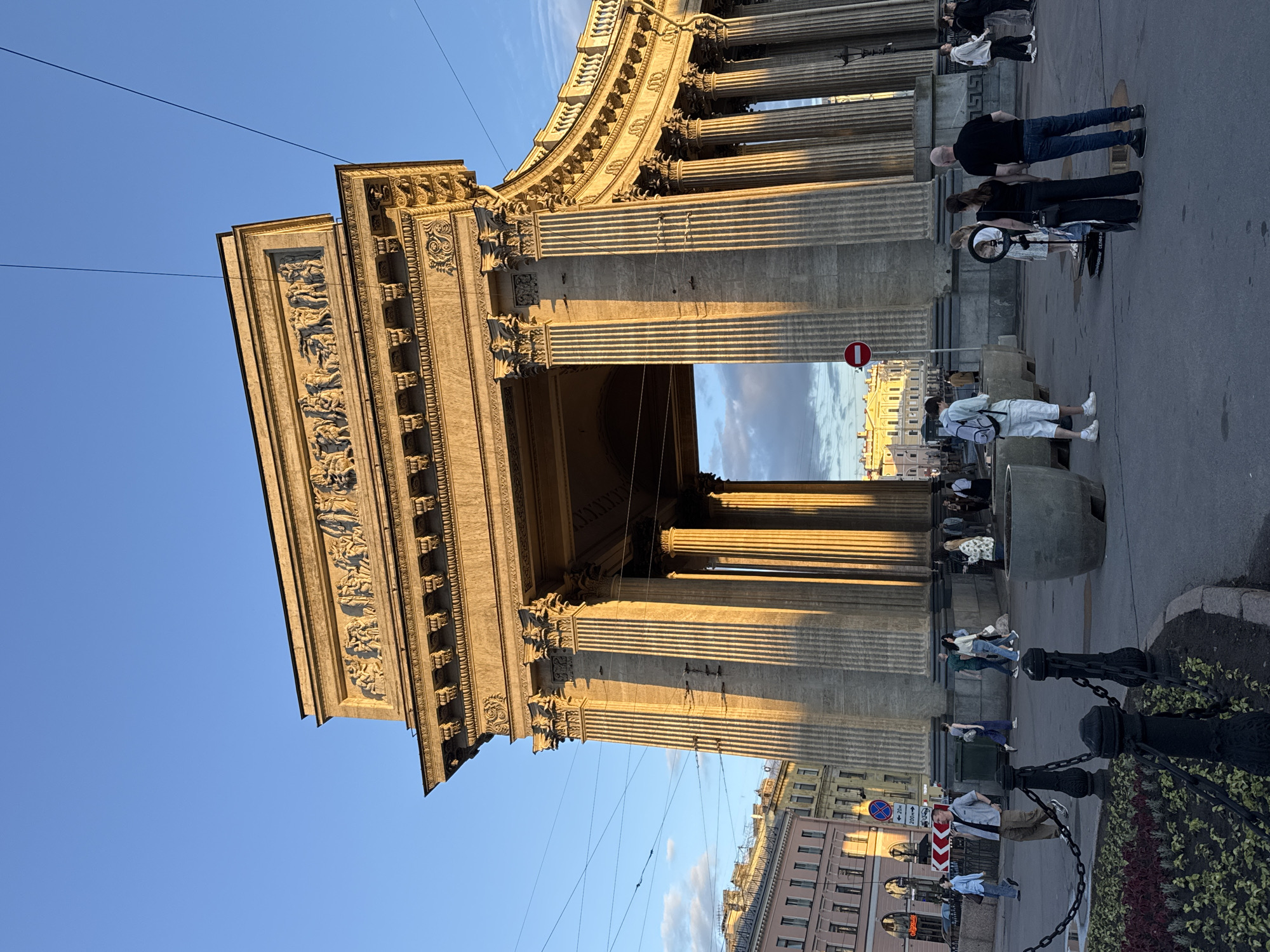 The image captures the Kazan Cathedral in Saint Petersburg, Russia, during what appears to be the golden hour of late afternoon. The majestic neoclassical colonnade of the cathedral forms the central focus, with its towering Corinthian columns casting long shadows. Above the portico, a relief frieze depicts classical figures. The sky is a clear, vibrant blue with a few wispy clouds.
In the foreground, a diverse group of people are casually walking and standing around the plaza. Some are dressed in casual attire, while others appear to be tourists. A few individuals are near a prominent circular sign with a red border, possibly indicating a no-entry zone. There are also large, weathered planters and decorative chains bordering a flowerbed. In the background, other historic buildings of Saint Petersburg can be seen, contributing to the urban landscape. The lighting suggests a warm, late afternoon sun, highlighting the architectural details of the cathedral and creating a serene yet lively atmosphere.